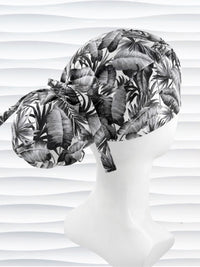 Ponytail style surgical scrub cap by JensCaps with tropical leaves in black and white on a soft white background cotton fabric, finished with an adjustable tie and displayed on a white mannequin head.