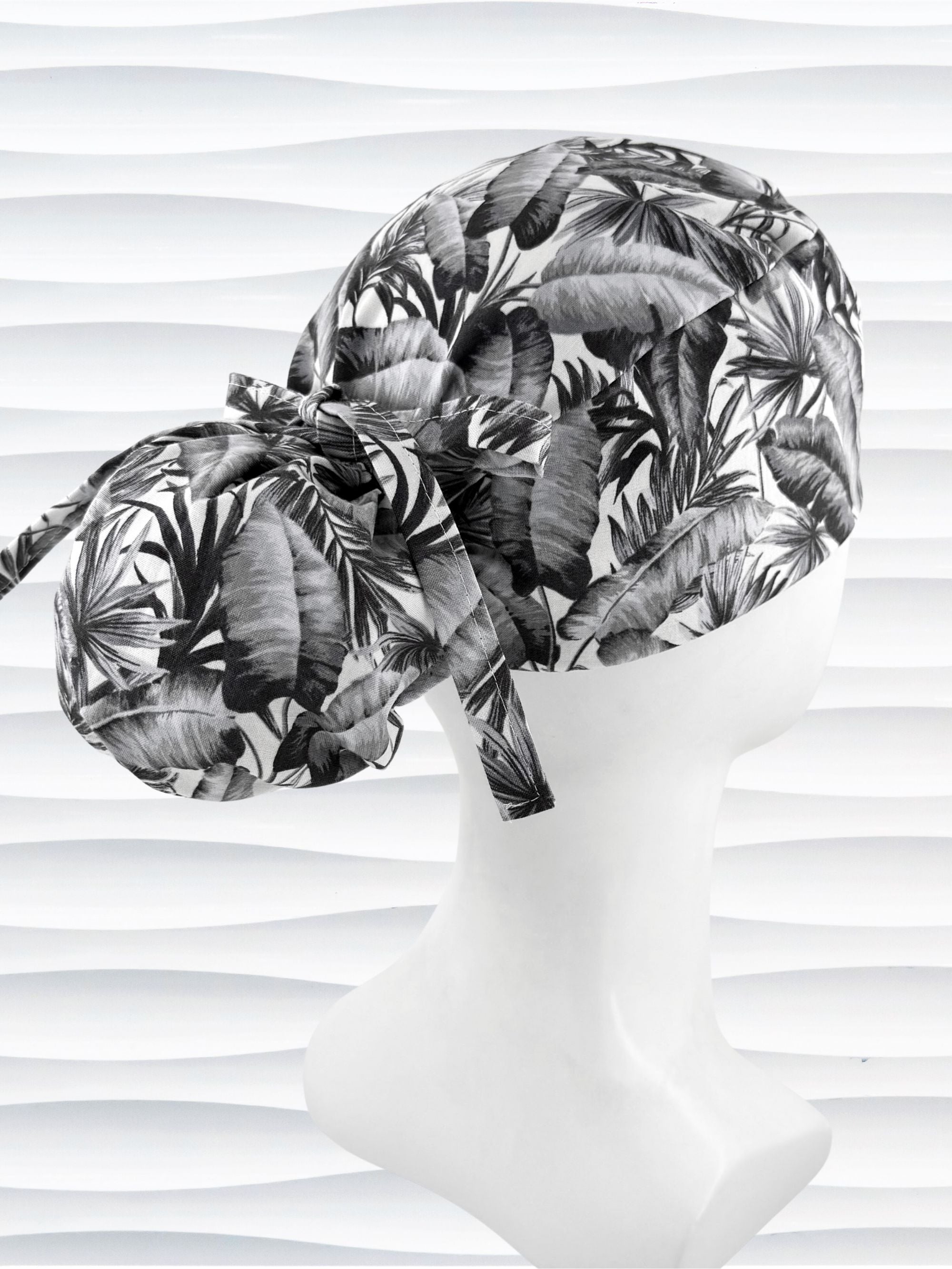 Ponytail style surgical scrub cap by JensCaps with tropical leaves in black and white on a soft white background cotton fabric, finished with an adjustable tie and displayed on a white mannequin head.