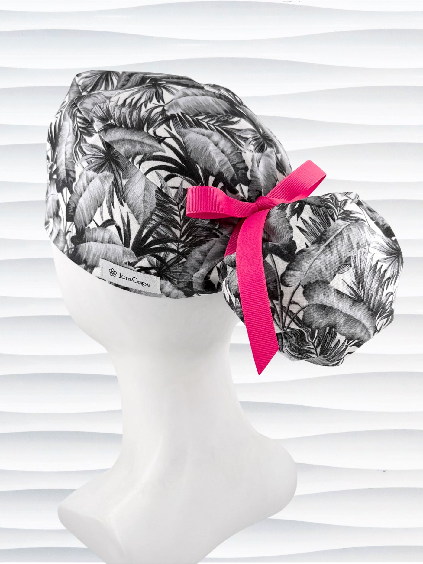Ponytail style surgical scrub cap by JensCaps with tropical leaves in black and white on a soft white background cotton fabric, finished with an adjustable tie and displayed on a white mannequin head.