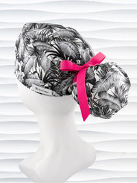 Ponytail style surgical scrub cap by JensCaps with tropical leaves in black and white on a soft white background cotton fabric, finished with an adjustable tie and displayed on a white mannequin head.