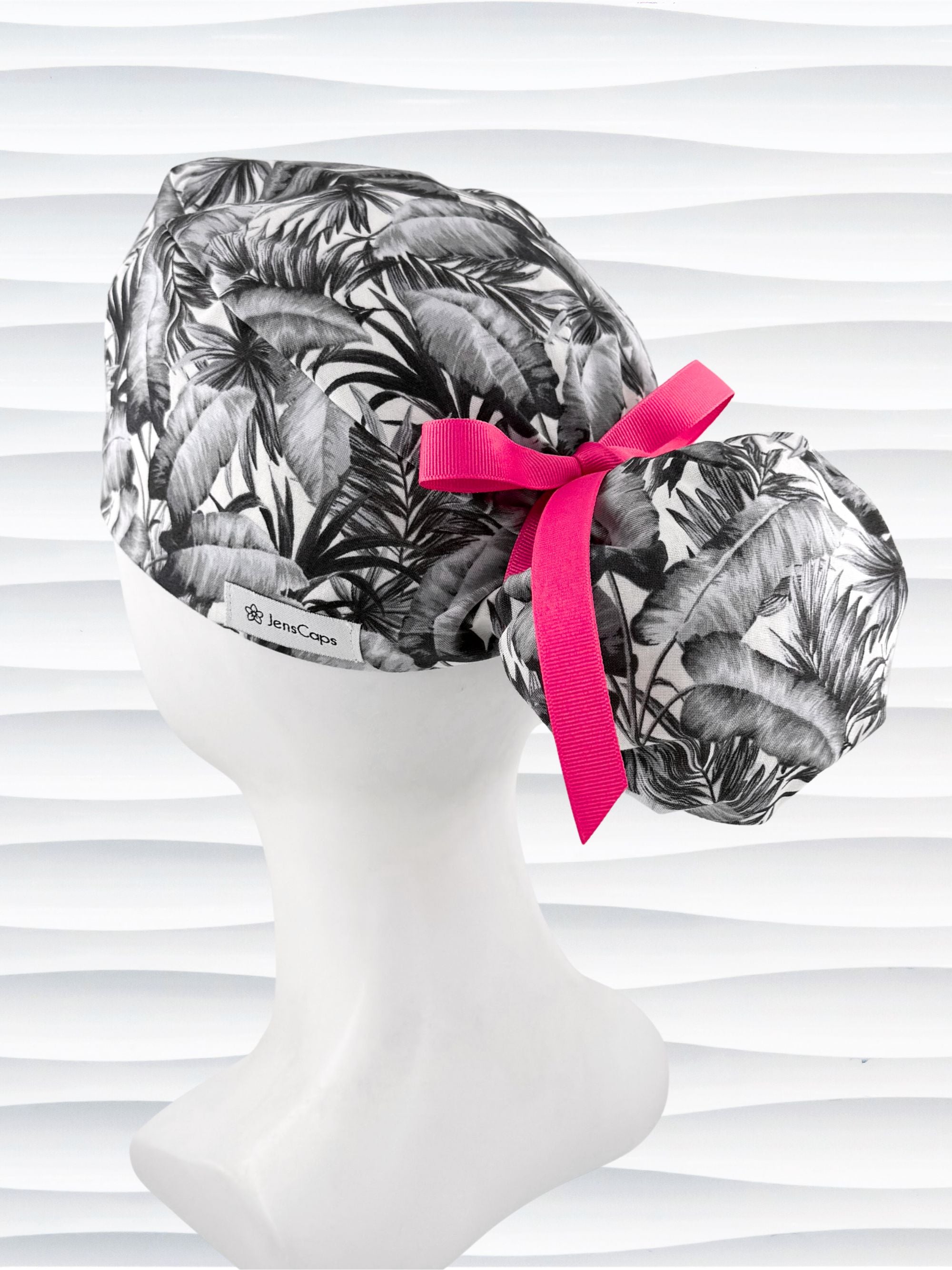 Ponytail style surgical scrub cap by JensCaps with tropical leaves in black and white on a soft white background cotton fabric, finished with an adjustable tie and displayed on a white mannequin head.