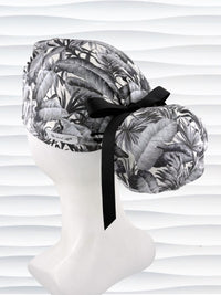 Ponytail style surgical scrub cap by JensCaps with tropical leaves in black and white on a soft white background cotton fabric, finished with an adjustable tie and displayed on a white mannequin head.