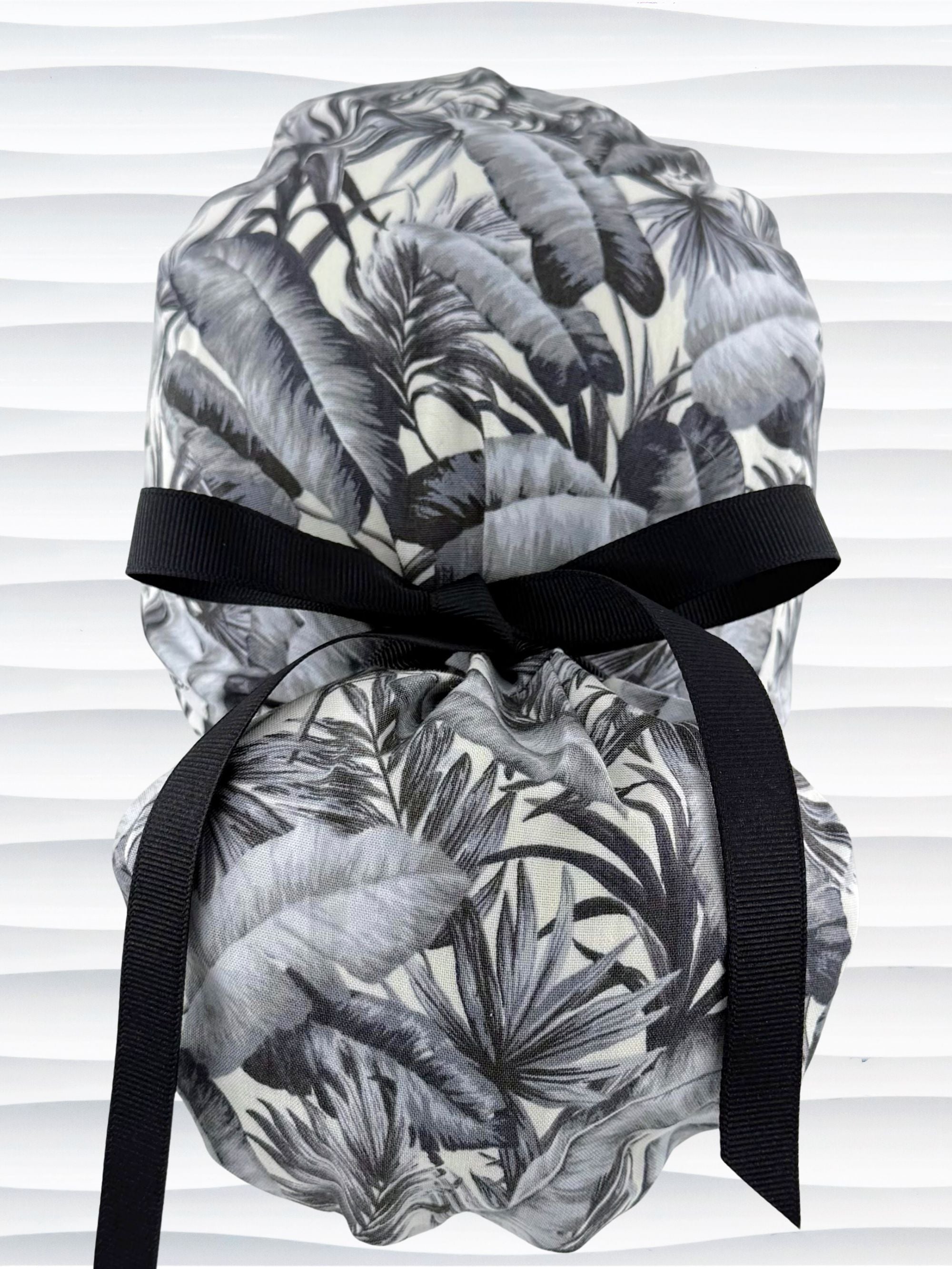 Ponytail style surgical scrub cap by JensCaps with tropical leaves in black and white on a soft white background cotton fabric, finished with an adjustable tie and displayed on a white mannequin head.