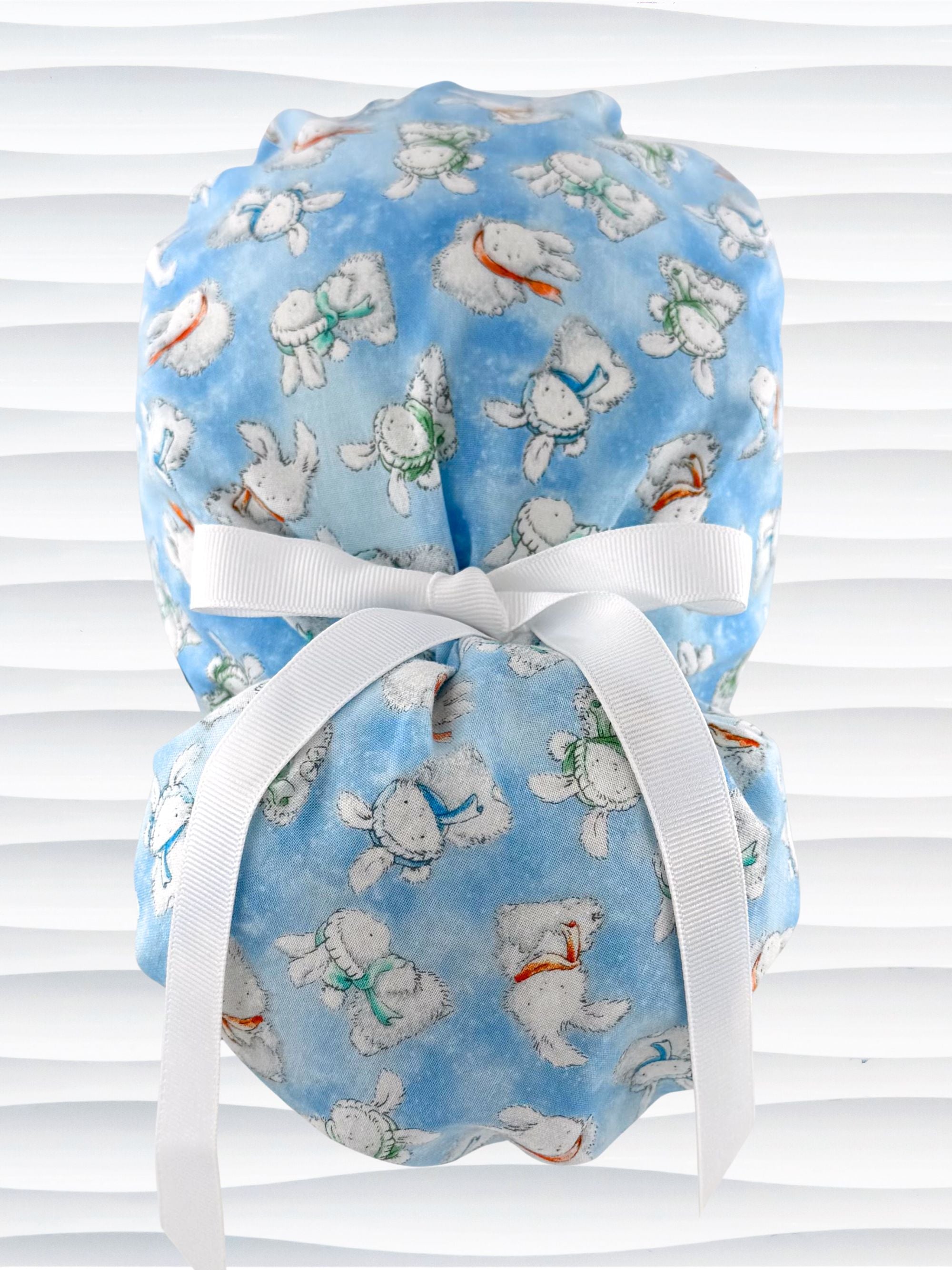 Ponytail style surgical scrub cap by JensCaps with white bunnies wearing beanies and scarves tossed all over this light blue cotton fabric, finished with a ribbon tie, and displayed on a white mannequin head
