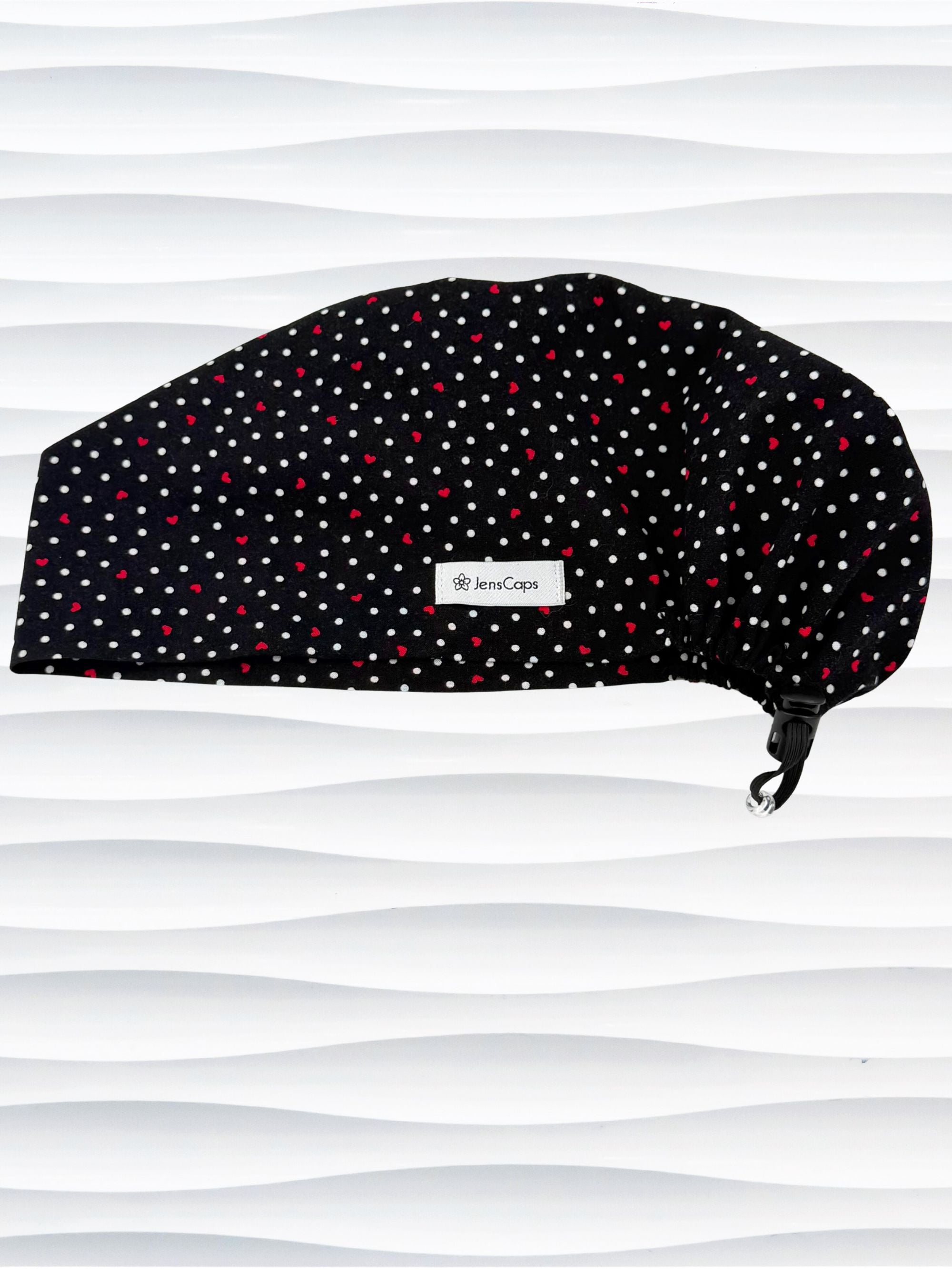Euro Pixie surgical scrub cap by JensCaps with small red hearts and white dots all over this black cotton fabric and displayed on a white mannequin head.