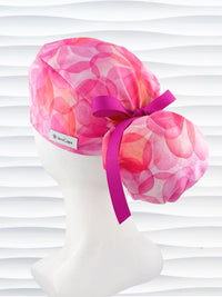 Ponytail  style surgical scrub cap by JensCaps with large flowers in light and dark pinks and peach sorbet on cotton fabric, finished with a ribbon tie, and displayed on a white mannequin head.