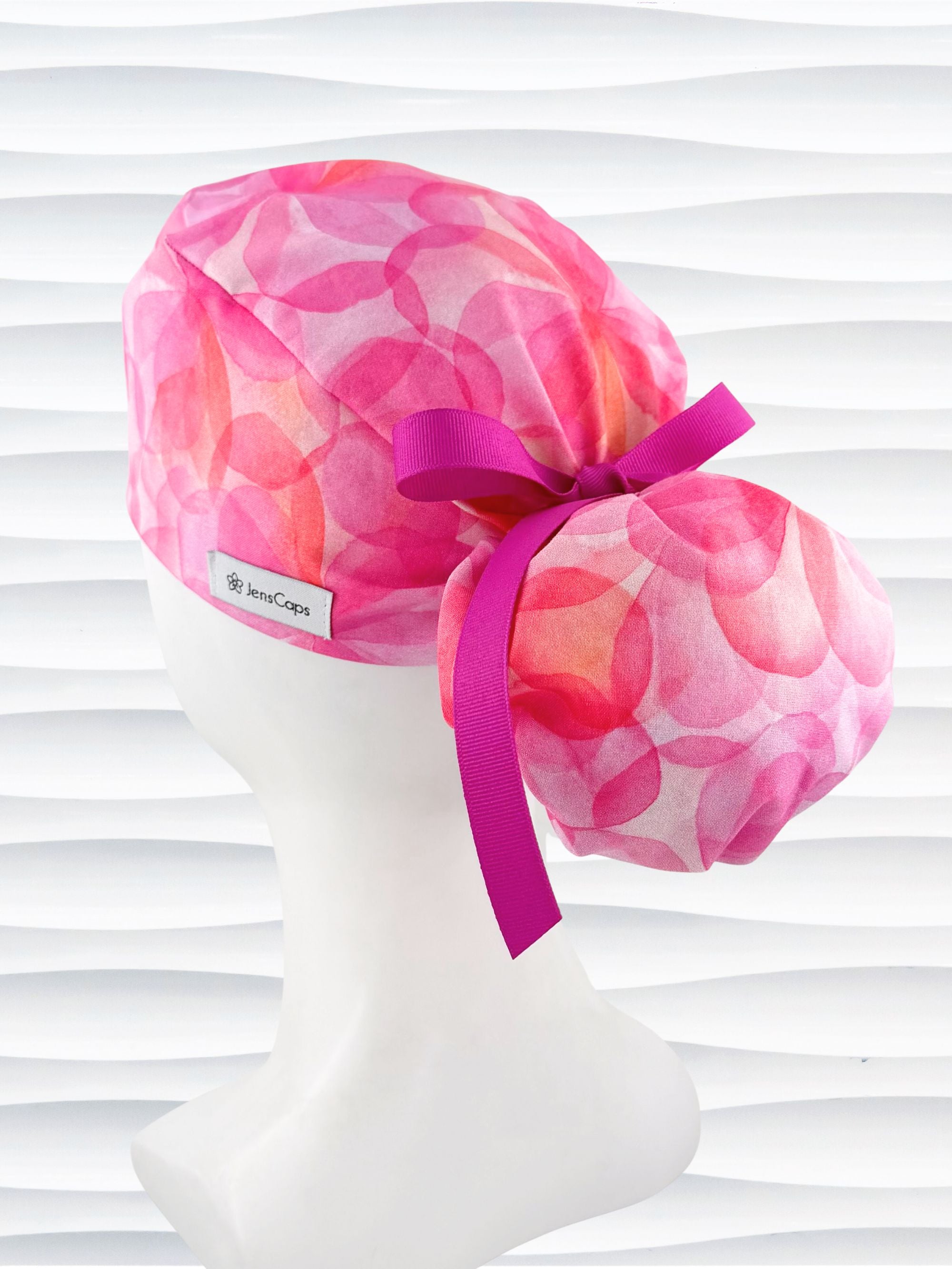 Ponytail  style surgical scrub cap by JensCaps with large flowers in light and dark pinks and peach sorbet on cotton fabric, finished with a ribbon tie, and displayed on a white mannequin head.