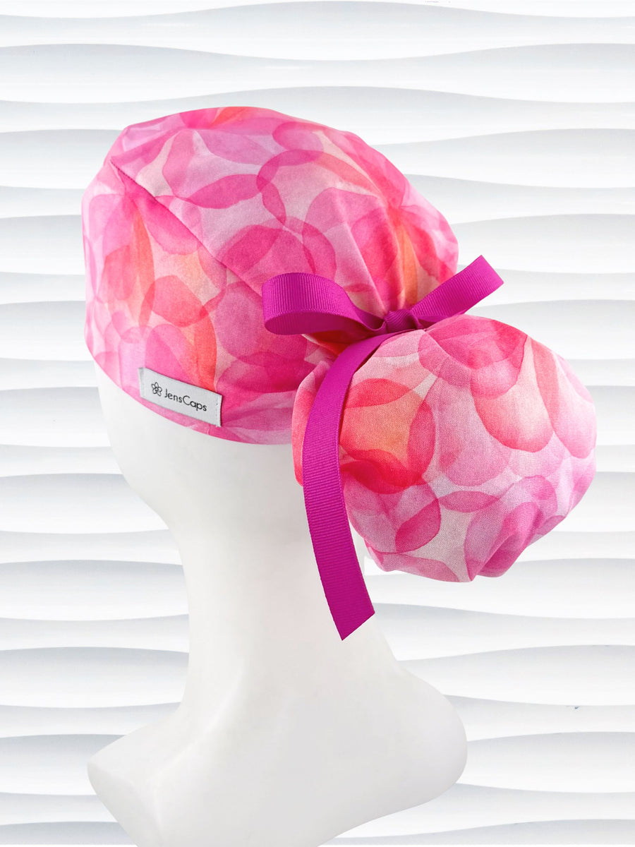 Ponytail  style surgical scrub cap by JensCaps with large flowers in light and dark pinks and peach sorbet on cotton fabric, finished with a ribbon tie, and displayed on a white mannequin head.