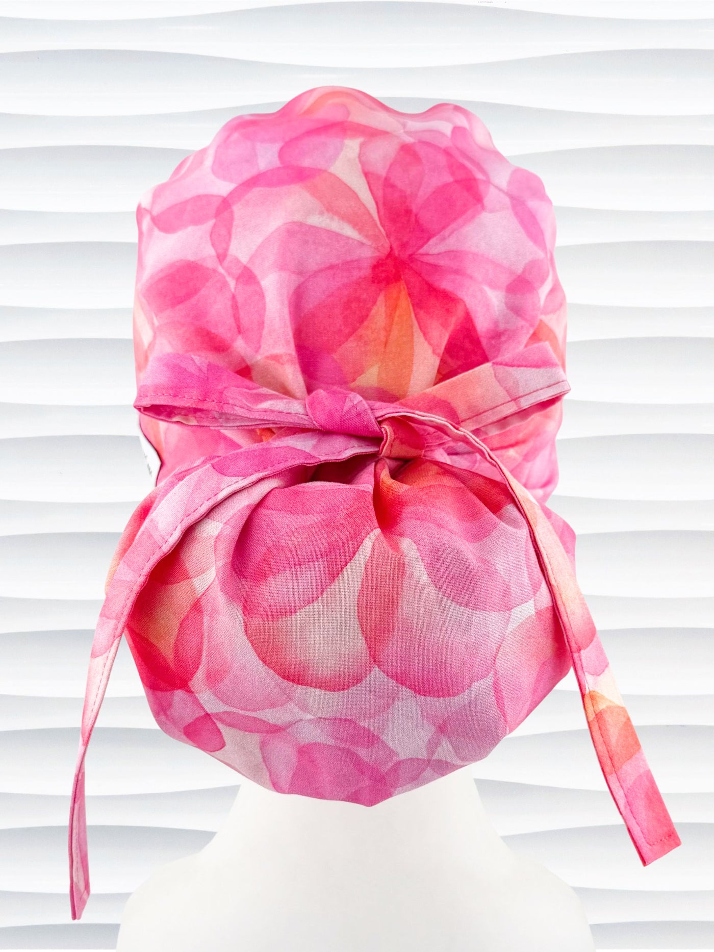 Ponytail  style surgical scrub cap by JensCaps with large flowers in light and dark pinks and peach sorbet on cotton fabric, finished with a ribbon tie, and displayed on a white mannequin head.