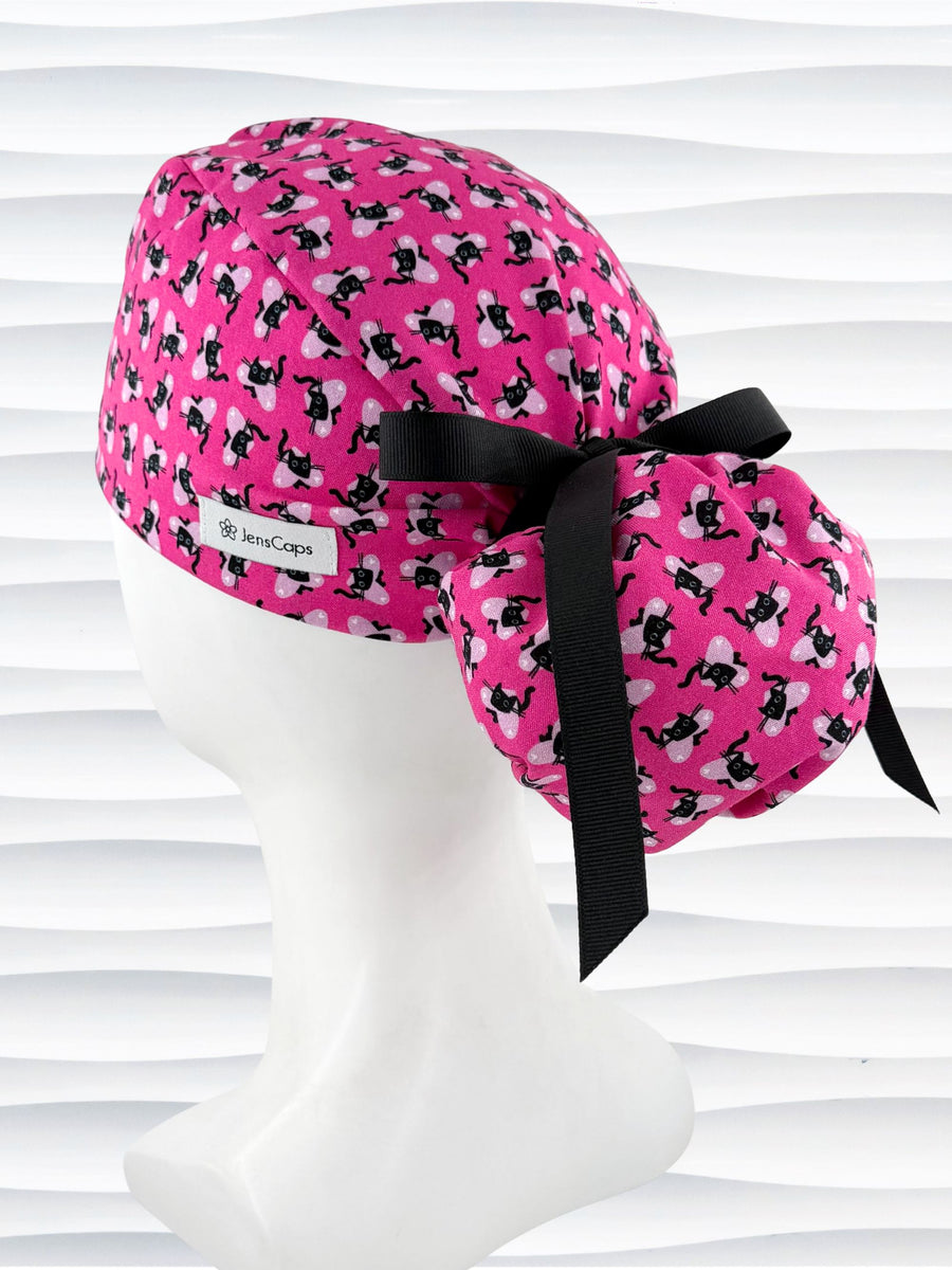Ponytail style surgical scrub cap by JensCaps with black cats and pink hearts all over this pink background cotton fabric, tied with a ribbon, and displayed on a white mannequin head.