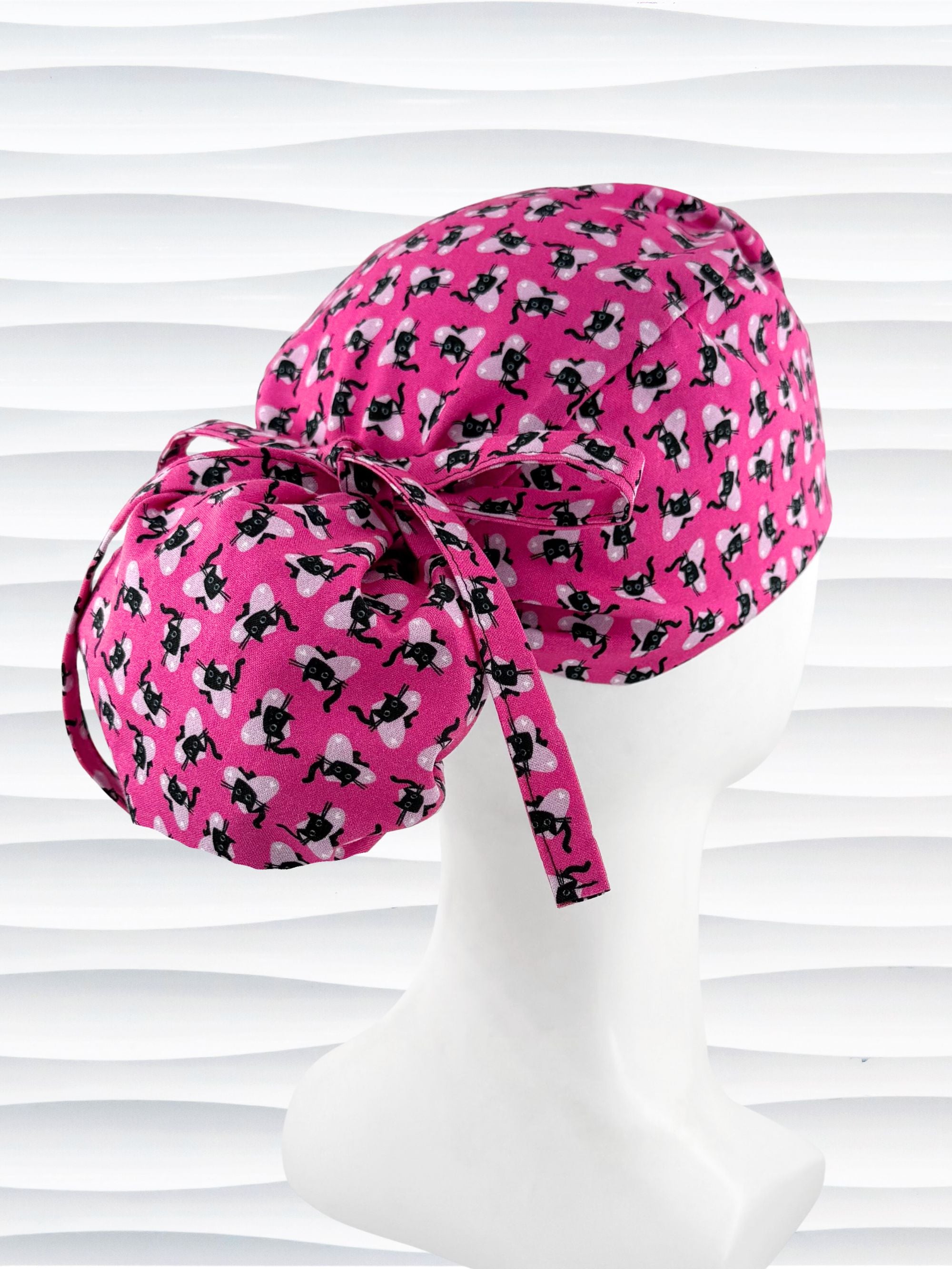 Ponytail style surgical scrub cap by JensCaps with black cats and pink hearts all over this pink background cotton fabric, tied with a ribbon, and displayed on a white mannequin head.