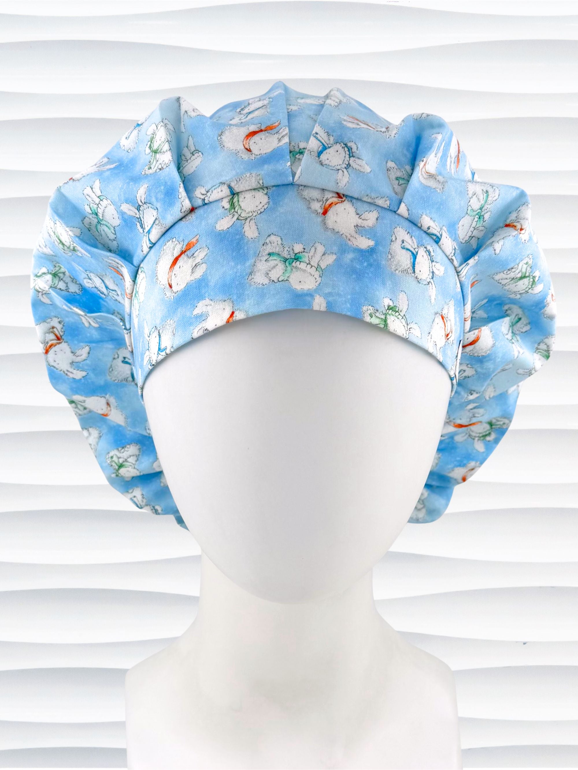Bouffant style surgical scrub cap by JensCaps with white bunnies wearing beanies and scarves tossed all over this light blue cotton fabric and displayed on a white mannequin head