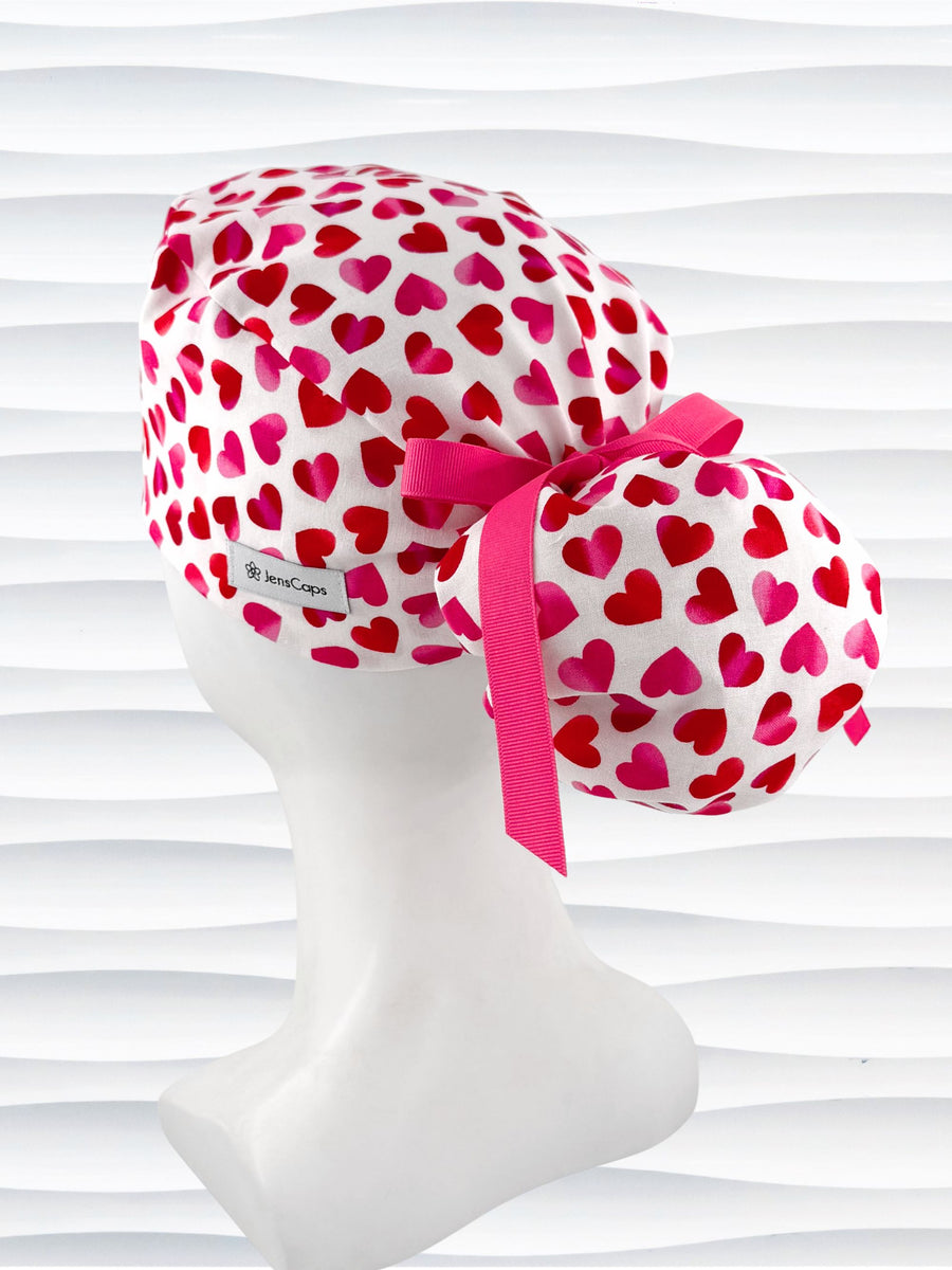 Ponytail style surgical scrub cap by JensCaps with pink to red ombre hearts all over a white background, finished with a ribbon tie, and displayed on a white mannequin head