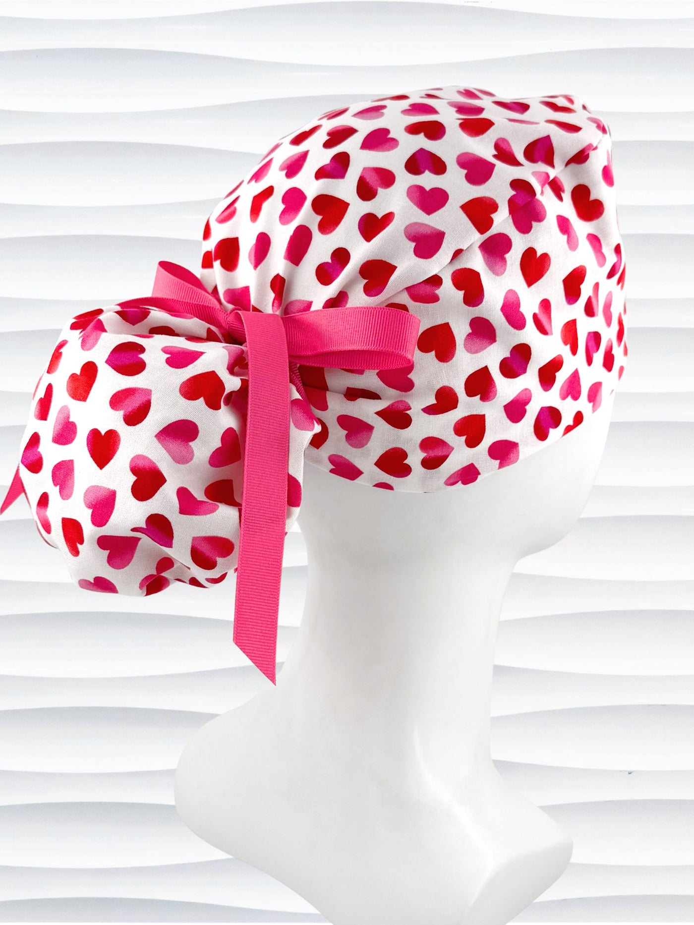 Ponytail style surgical scrub cap by JensCaps with pink to red ombre hearts all over a white background, finished with a ribbon tie, and displayed on a white mannequin head