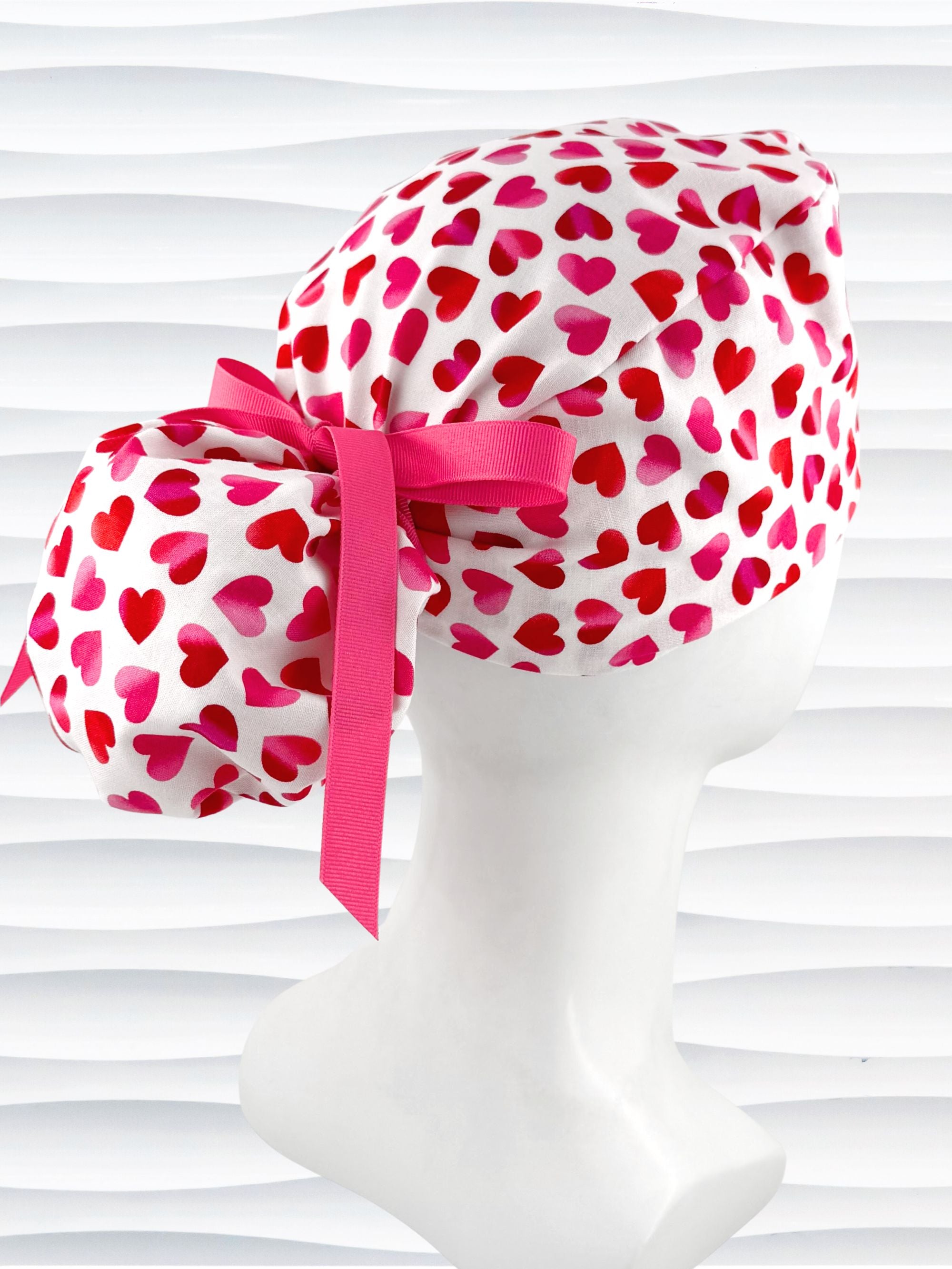 Ponytail style surgical scrub cap by JensCaps with pink to red ombre hearts all over a white background, finished with a ribbon tie, and displayed on a white mannequin head