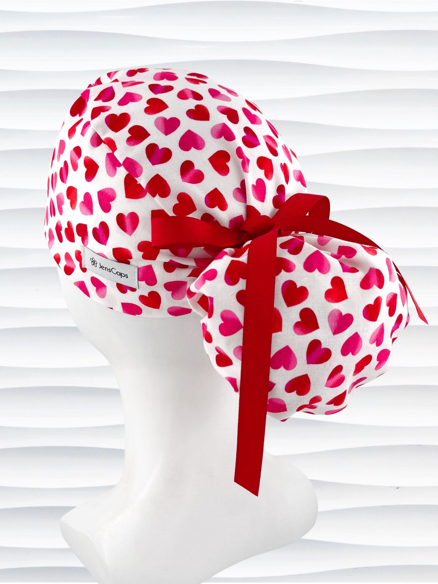 Ponytail style surgical scrub cap by JensCaps with pink to red ombre hearts all over a white background, finished with a ribbon tie, and displayed on a white mannequin head