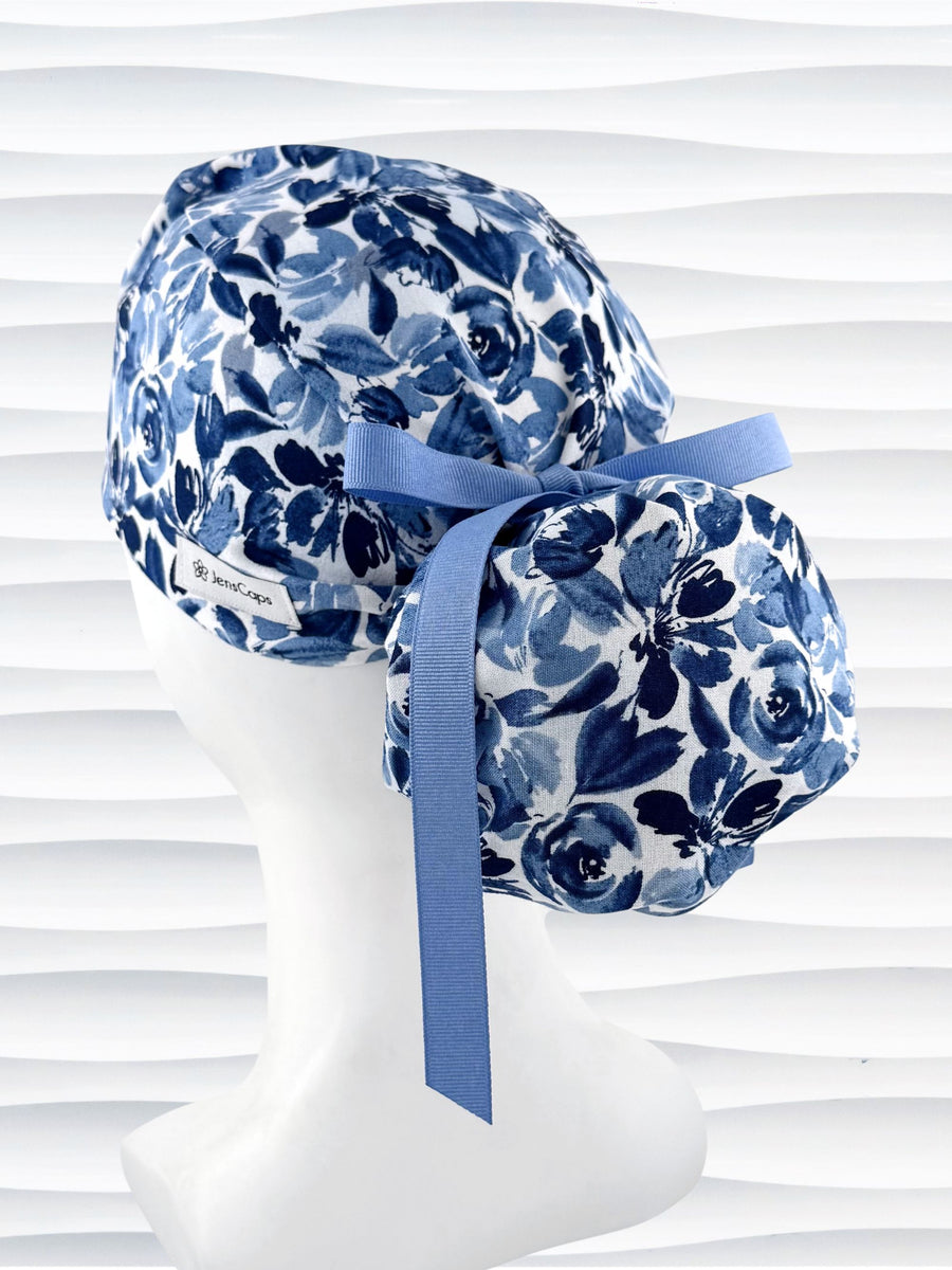 Ponytail style surgical scrub cap by JensCaps with flowers in varying shades of blue with a white background, finished with a ribbon tie, and displayed on a white mannequin head.