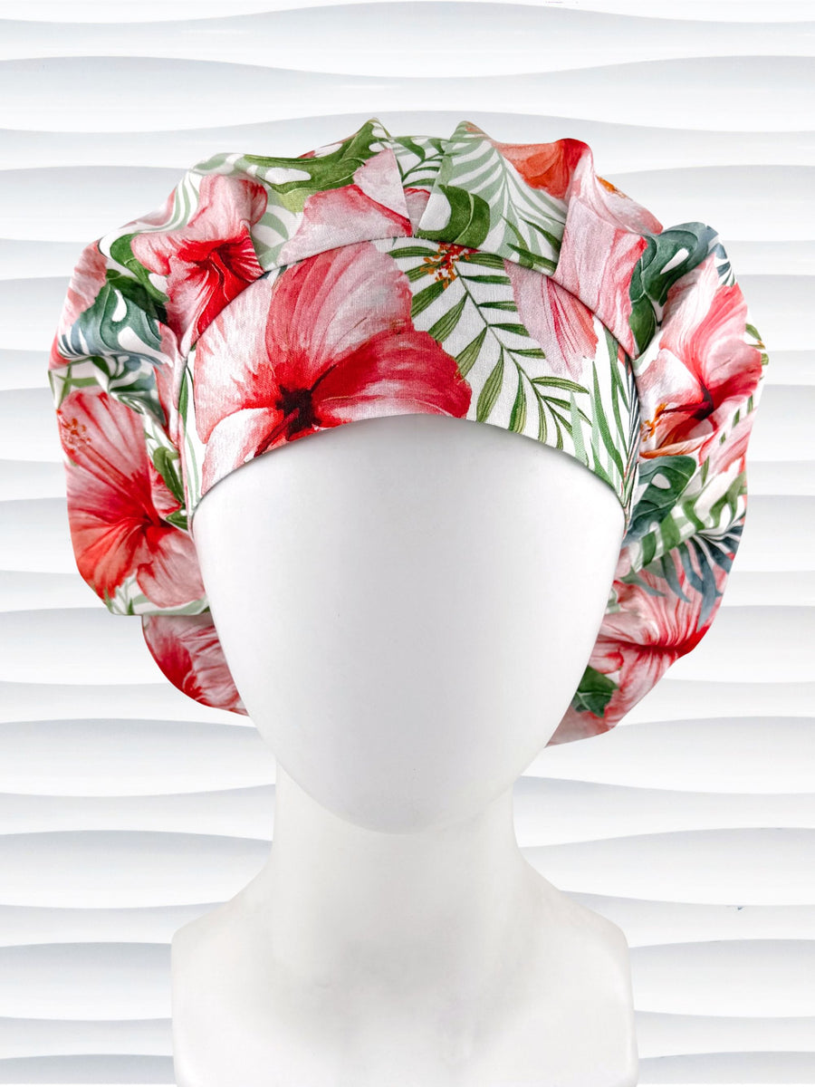 Bouffant style surgical scrub hat with red hibiscus flowers and green palm fronds all over white cotton fabric and displayed on a mannequin head.