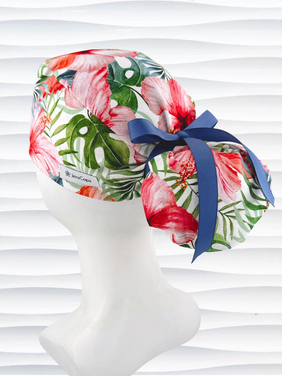 Ponytail style surgical scrub cap by JensCaps with tropical hibiscus flowers and palm fronds all over white cotton fabric, finished with a grosgrain ribbon tie, and displayed on a mannequin head