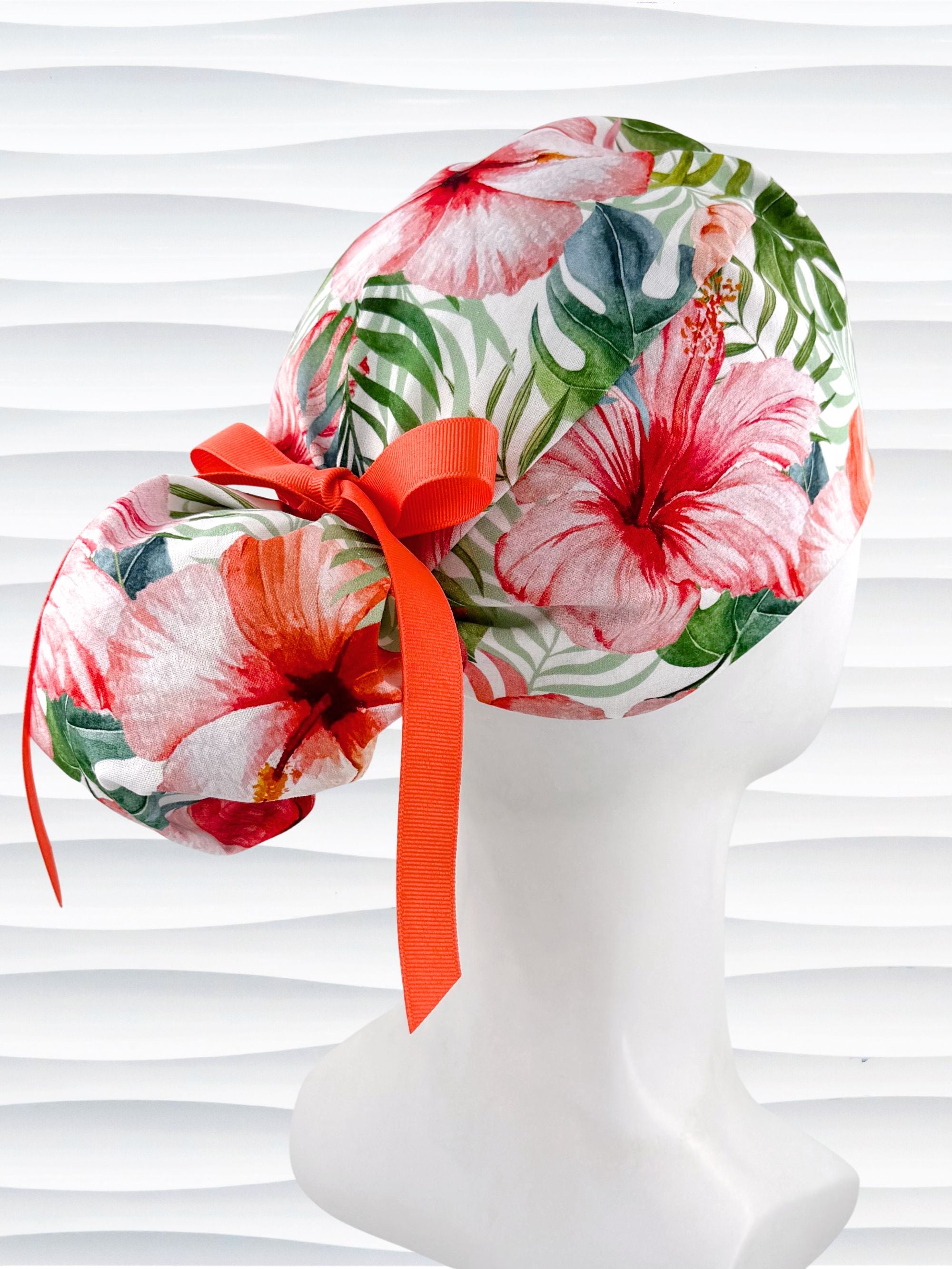 Ponytail style surgical scrub cap by JensCaps with tropical hibiscus flowers and palm fronds all over white cotton fabric, finished with a grosgrain ribbon tie, and displayed on a mannequin head