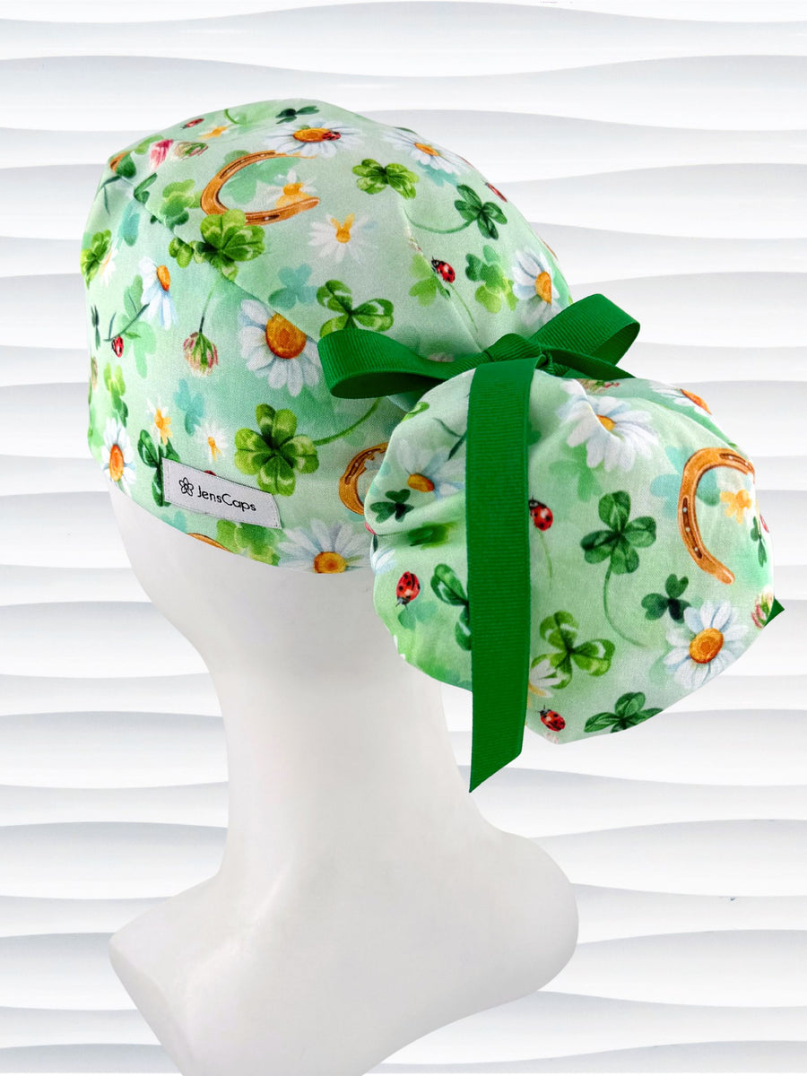 Ponytail style surgical scrub cap by JensCaps with clover, daisies, gold horseshoes, and 4 leaf clovers all over light green cotton fabric, finished with a grosgrain ribbon, and displayed on a mannequin head.