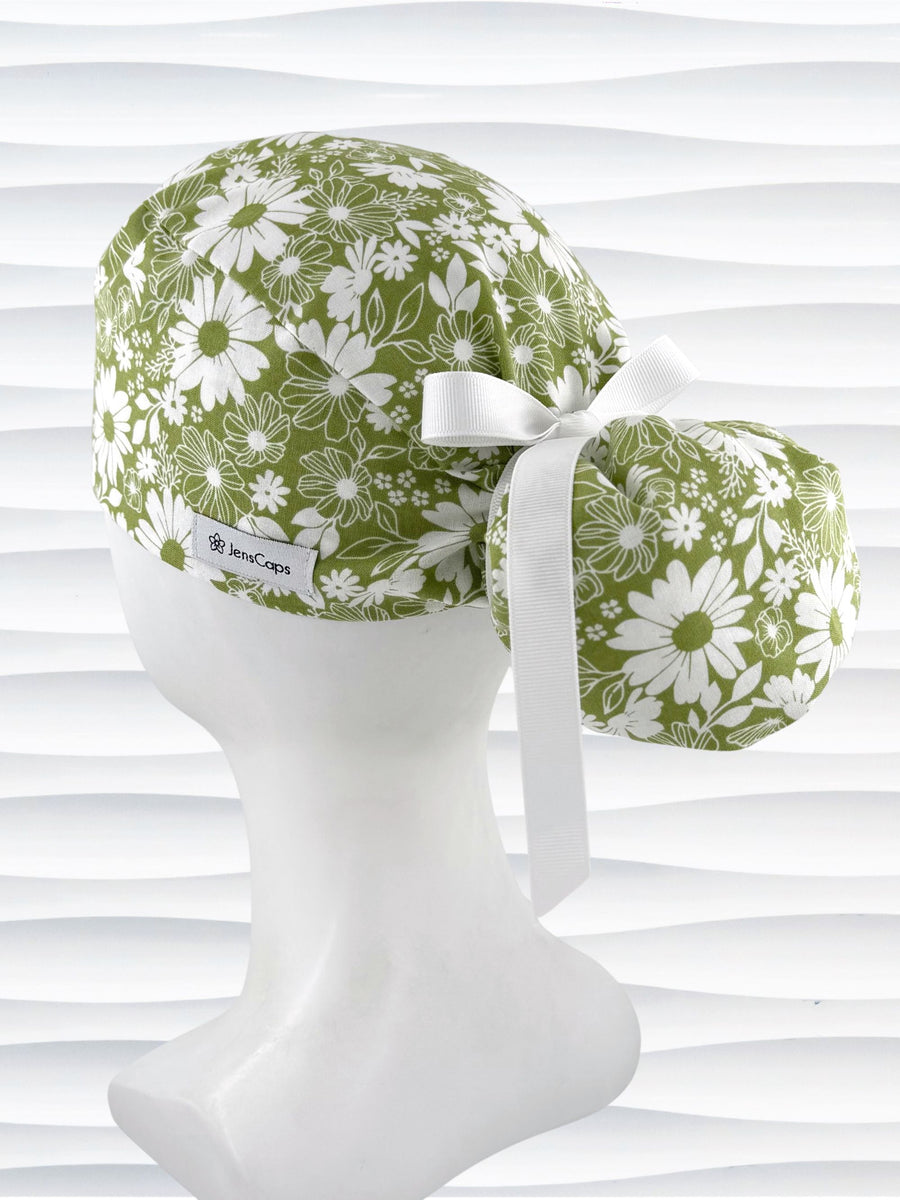 Ponytail style surgical scrub cap with white daisies and flowers all over a light green cotton fabric, finished with a grosgrain ribbon tie, and displayed on a mannequin head.