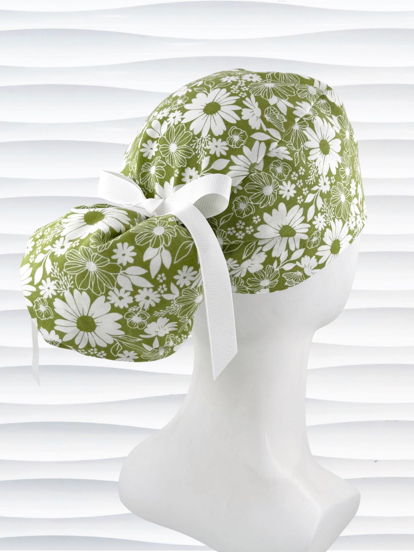 Ponytail style surgical scrub cap with white daisies and flowers all over a light green cotton fabric, finished with a grosgrain ribbon tie, and displayed on a mannequin head.