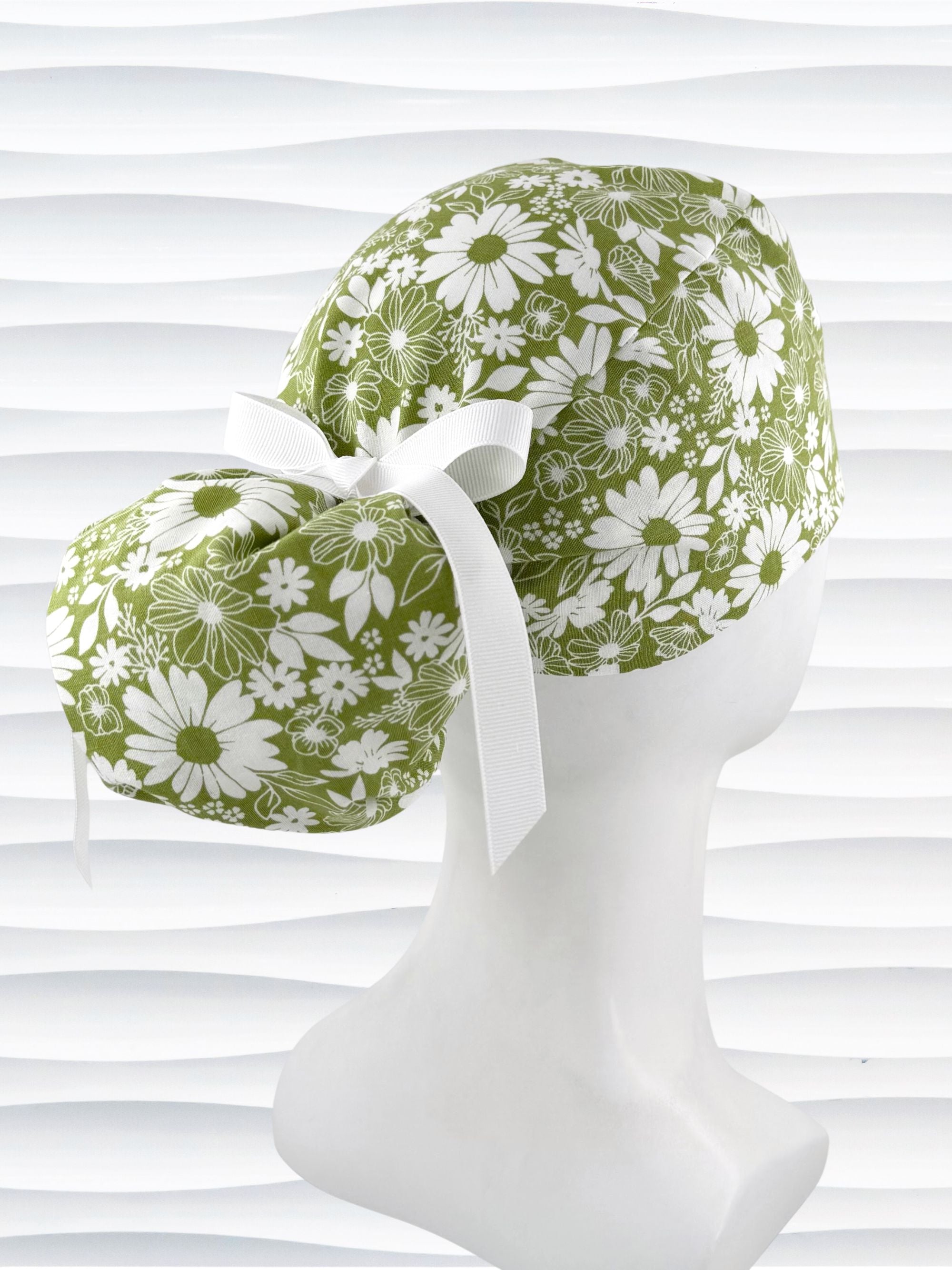 Ponytail style surgical scrub cap with white daisies and flowers all over a light green cotton fabric, finished with a grosgrain ribbon tie, and displayed on a mannequin head.
