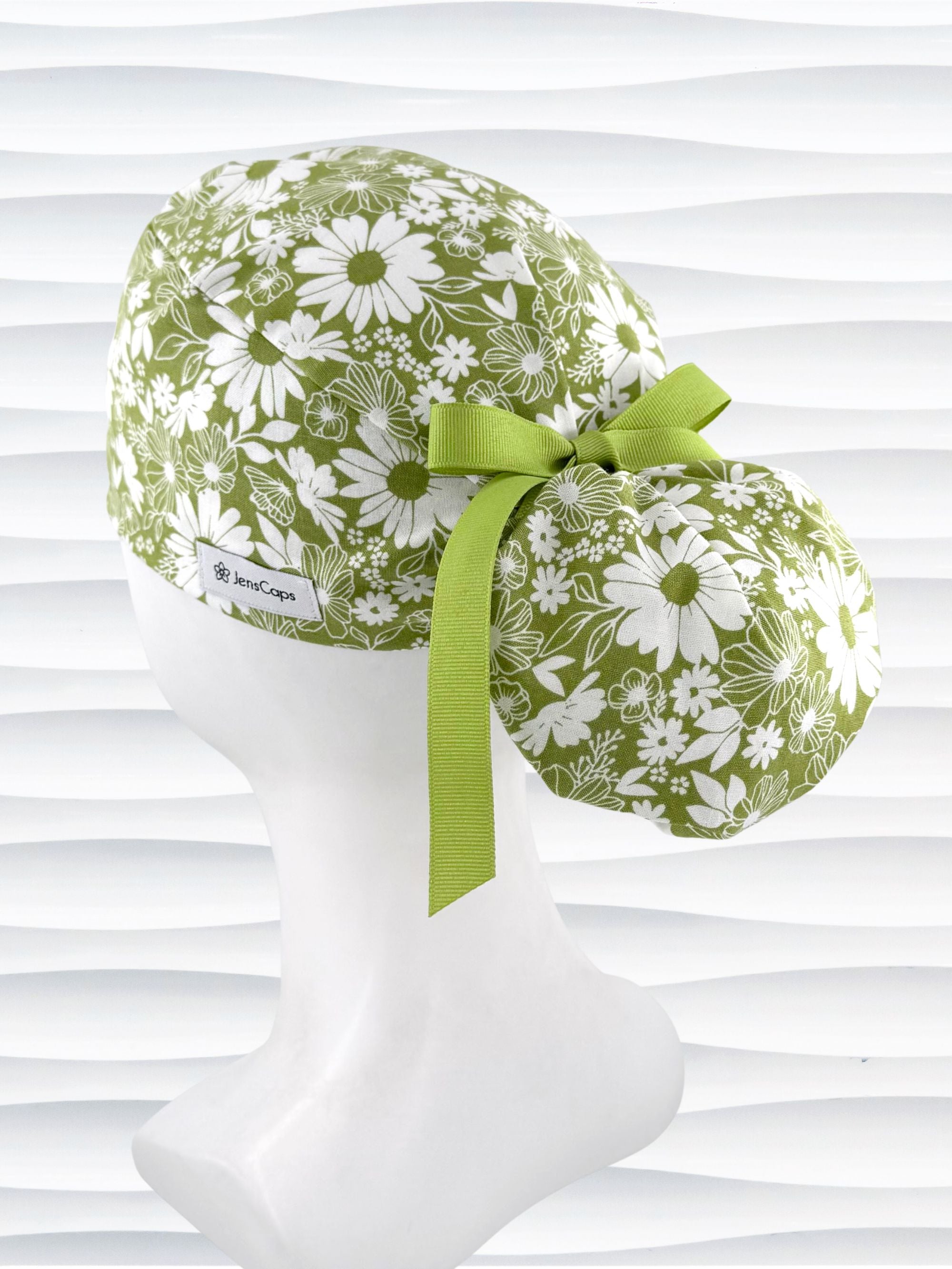 Ponytail style surgical scrub cap with white daisies and flowers all over a light green cotton fabric, finished with a grosgrain ribbon tie, and displayed on a mannequin head.