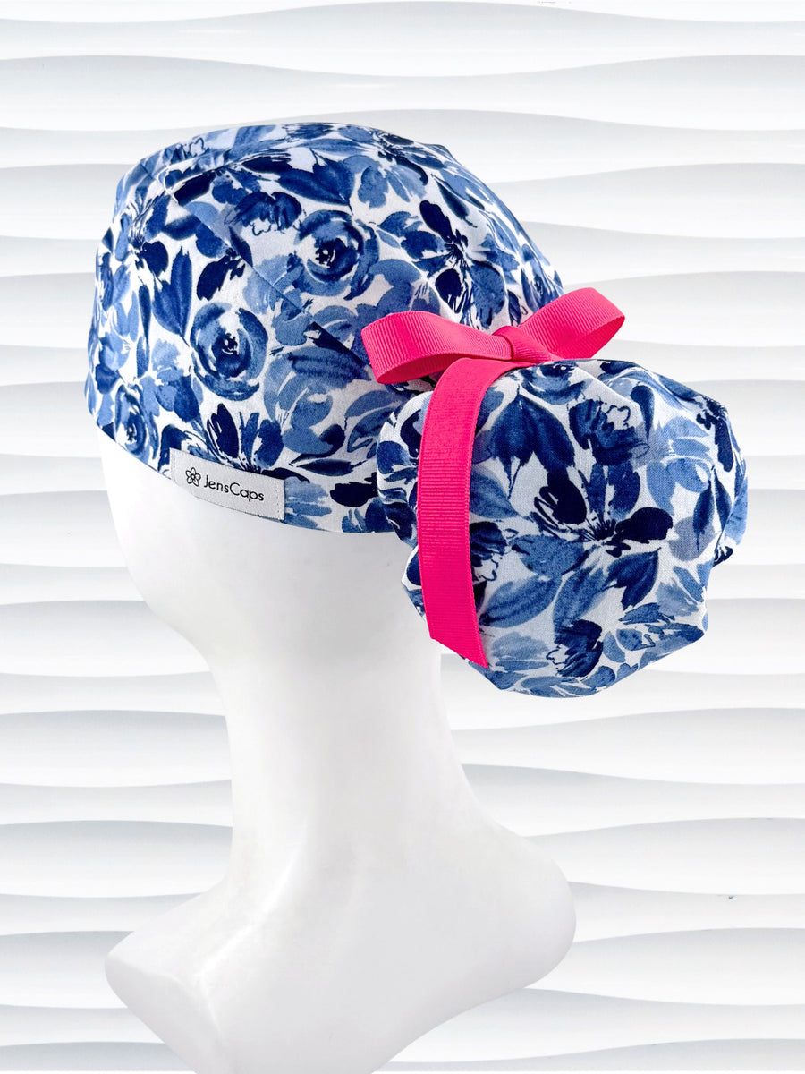 Ponytail style surgical scrub cap by JensCaps with flowers in varying shades of blue with a white background, finished with a ribbon tie, and displayed on a white mannequin head.