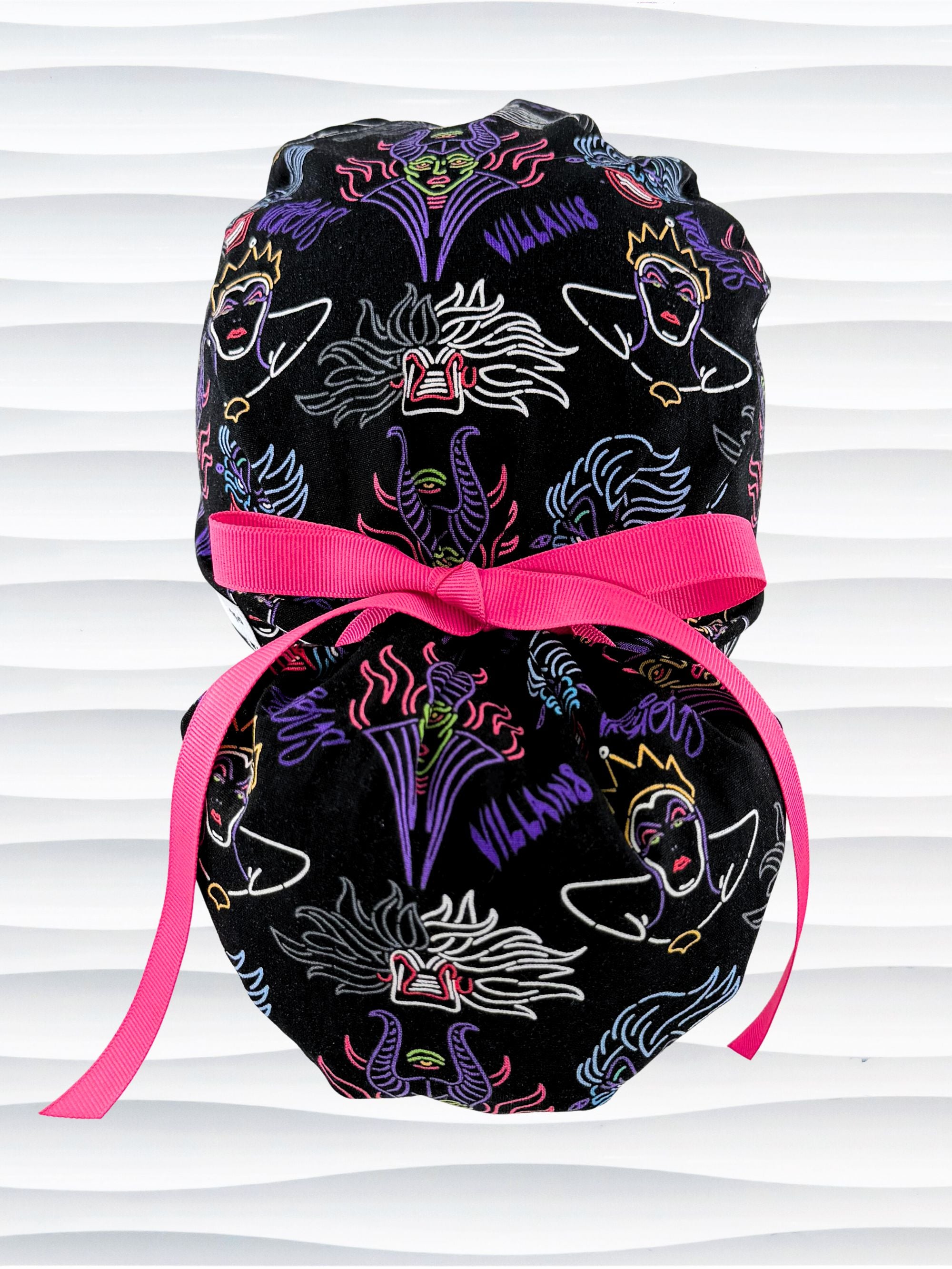 Ponytail style surgical scrub cap with neon colors of iconic villains all over black cotton fabric, finished with a ribbon tie and displayed on mannequin head.