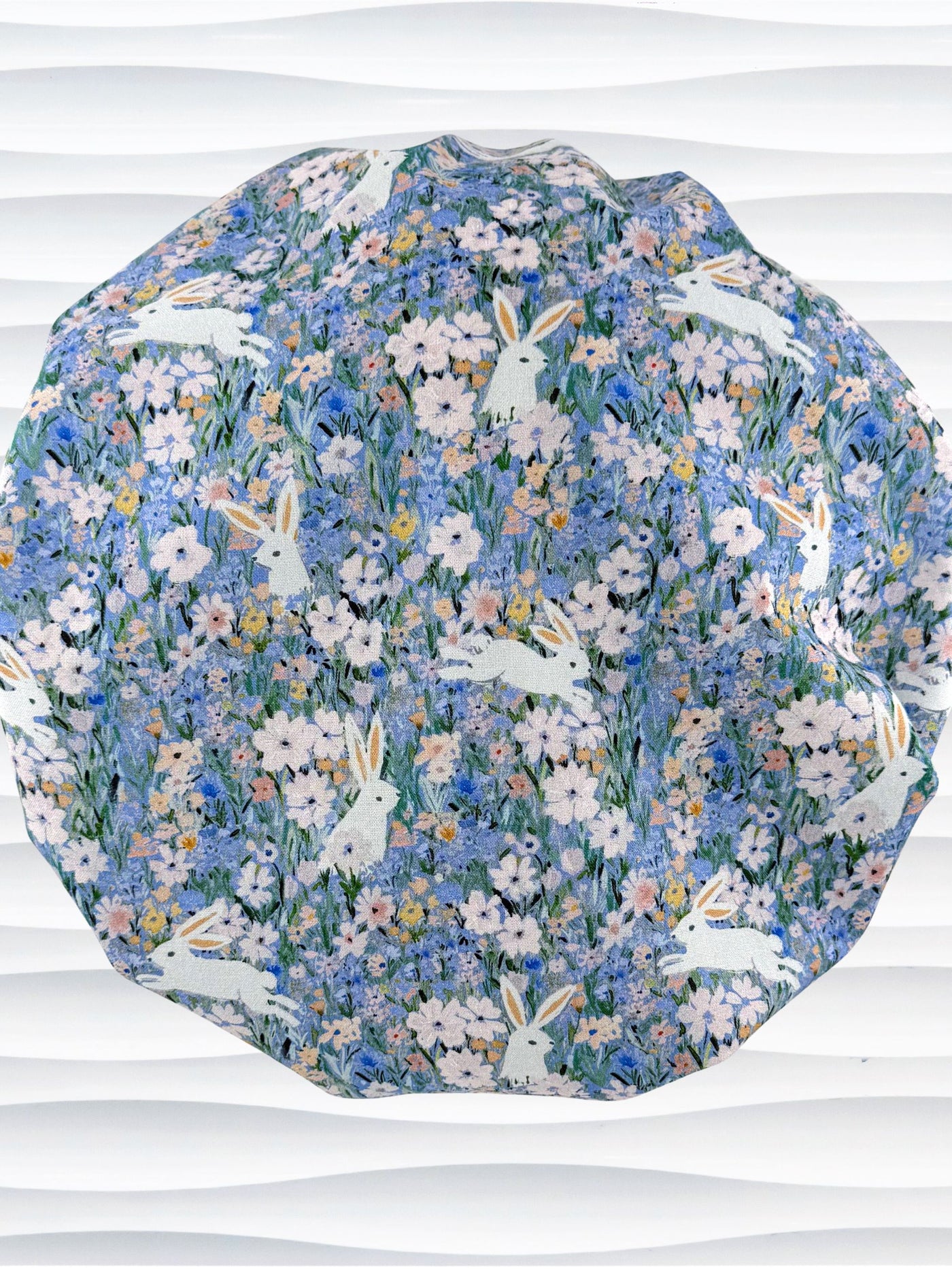 A bouffant style surgical scrub cap by JensCaps with white rabbits in various poses — sitting, leaping, and hopping — scattered across a dense meadow of wildflowers in periwinkle blue, soft pink, pale yellow, and green and displayed on a mannequin head.