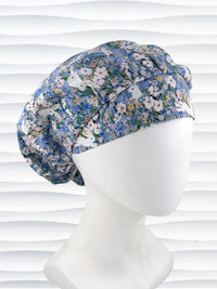 A bouffant style surgical scrub cap by JensCaps with white rabbits in various poses — sitting, leaping, and hopping — scattered across a dense meadow of wildflowers in periwinkle blue, soft pink, pale yellow, and green and displayed on a mannequin head.