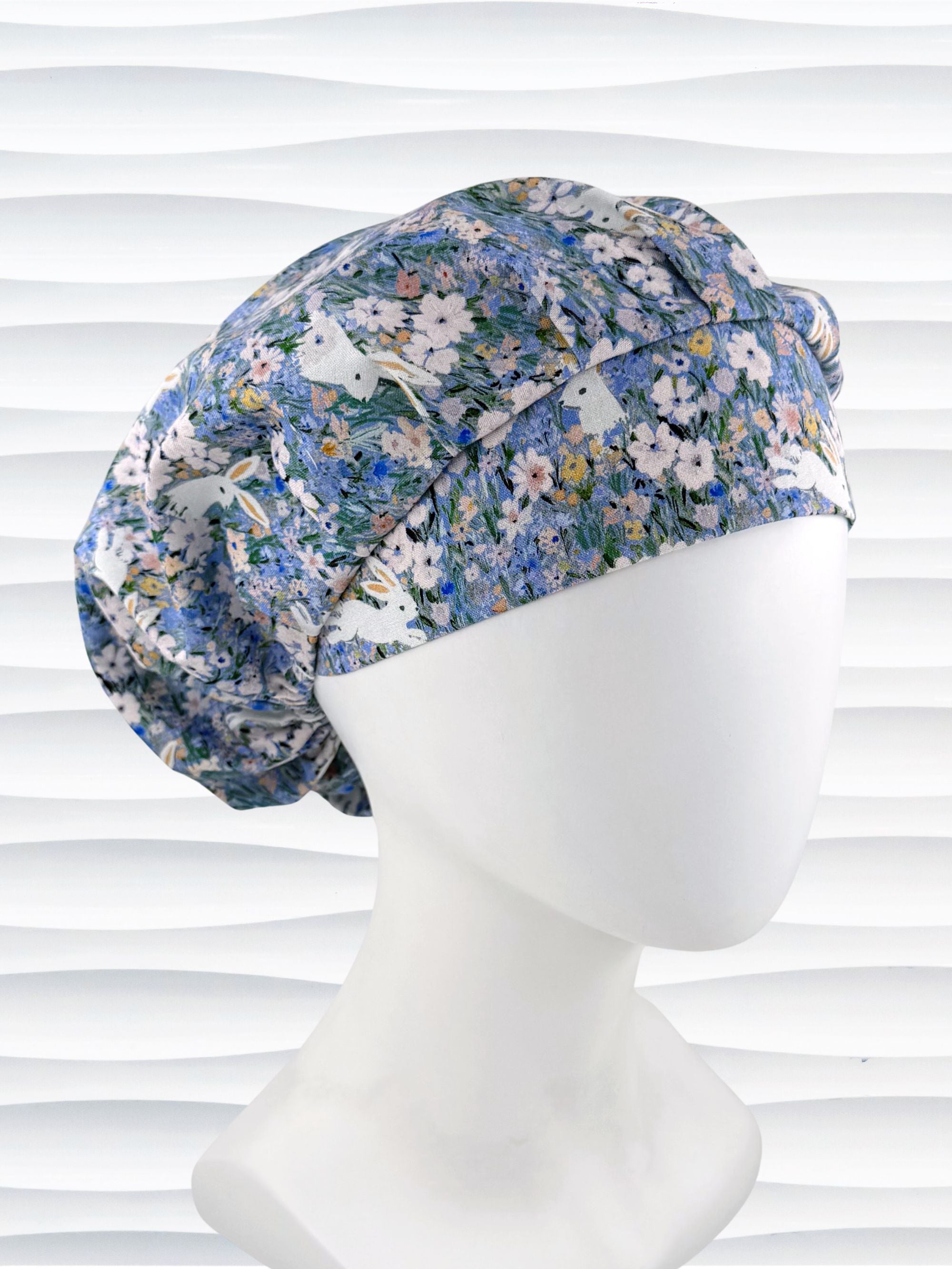 A bouffant style surgical scrub cap by JensCaps with white rabbits in various poses — sitting, leaping, and hopping — scattered across a dense meadow of wildflowers in periwinkle blue, soft pink, pale yellow, and green and displayed on a mannequin head.