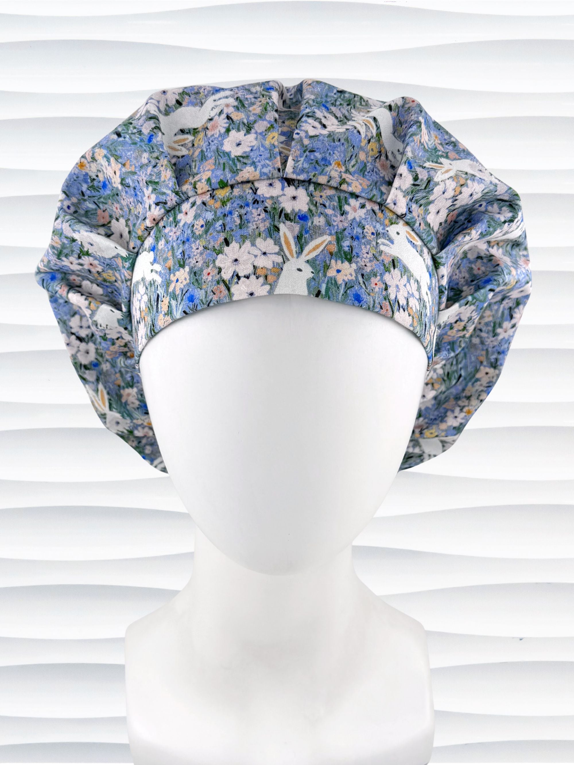 A bouffant style surgical scrub cap by JensCaps with white rabbits in various poses — sitting, leaping, and hopping — scattered across a dense meadow of wildflowers in periwinkle blue, soft pink, pale yellow, and green and displayed on a mannequin head.