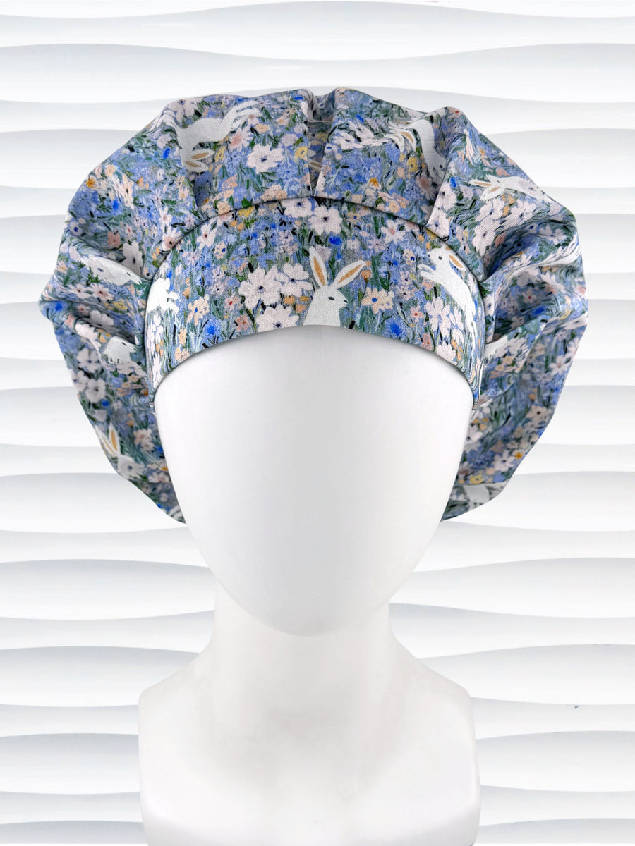 A bouffant style surgical scrub cap by JensCaps with white rabbits in various poses — sitting, leaping, and hopping — scattered across a dense meadow of wildflowers in periwinkle blue, soft pink, pale yellow, and green and displayed on a mannequin head.