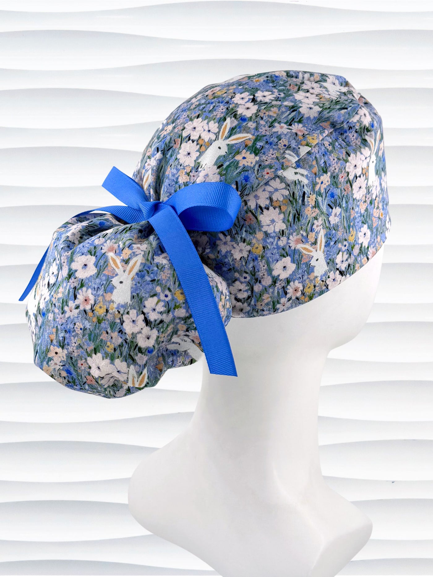 A ponytail style surgical scrub cap by JensCaps with white rabbits in various poses — sitting, leaping, and hopping — scattered across a dense meadow of wildflowers in periwinkle blue, soft pink, pale yellow, and green and finished with a ribbon tie and photographed on a mannequin head.