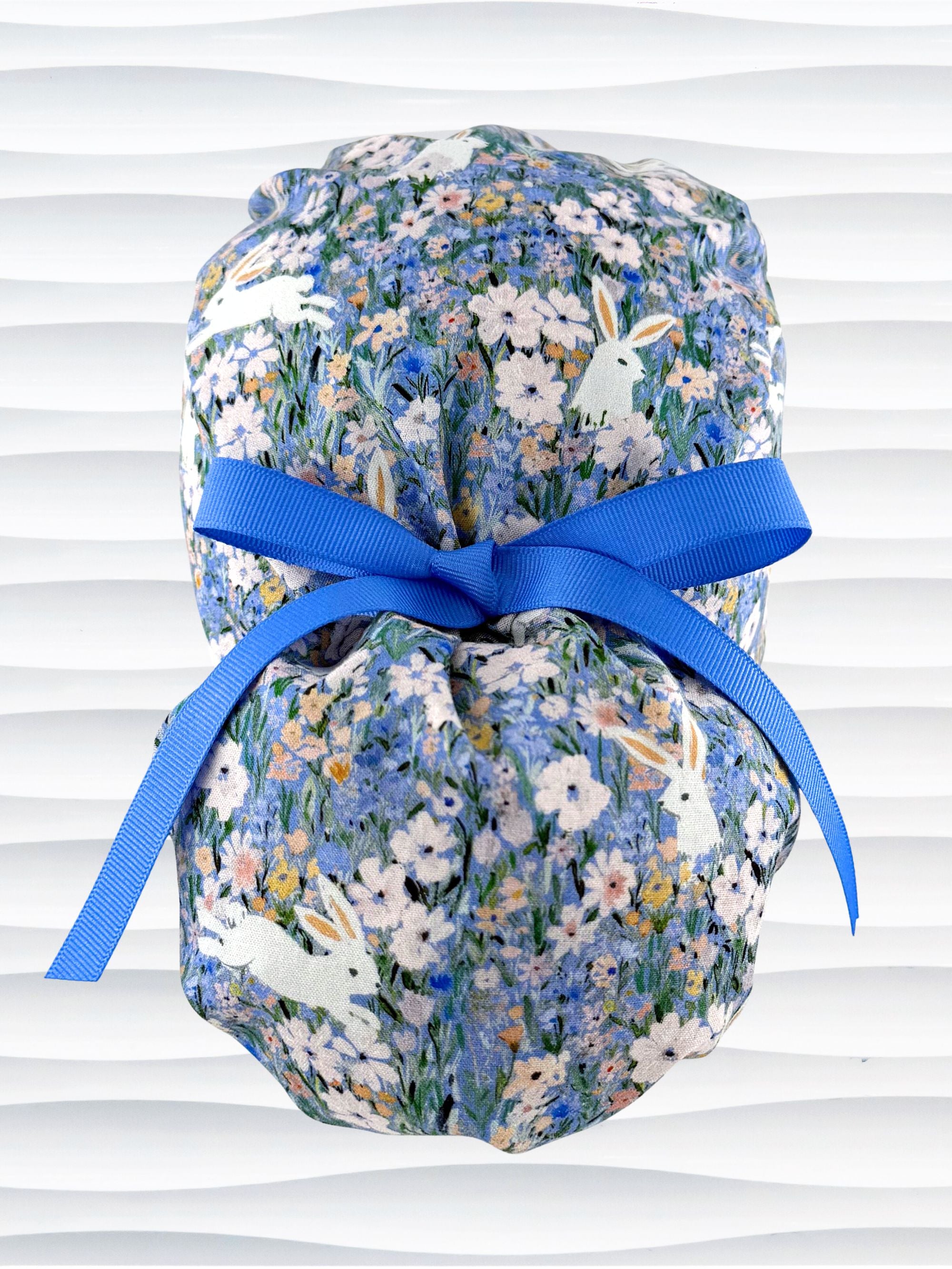 A ponytail style surgical scrub cap by JensCaps with white rabbits in various poses — sitting, leaping, and hopping — scattered across a dense meadow of wildflowers in periwinkle blue, soft pink, pale yellow, and green and finished with a ribbon tie and photographed on a mannequin head.