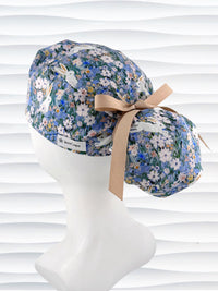 A ponytail style surgical scrub cap by JensCaps with white rabbits in various poses — sitting, leaping, and hopping — scattered across a dense meadow of wildflowers in periwinkle blue, soft pink, pale yellow, and green and finished with a ribbon tie and photographed on a mannequin head.