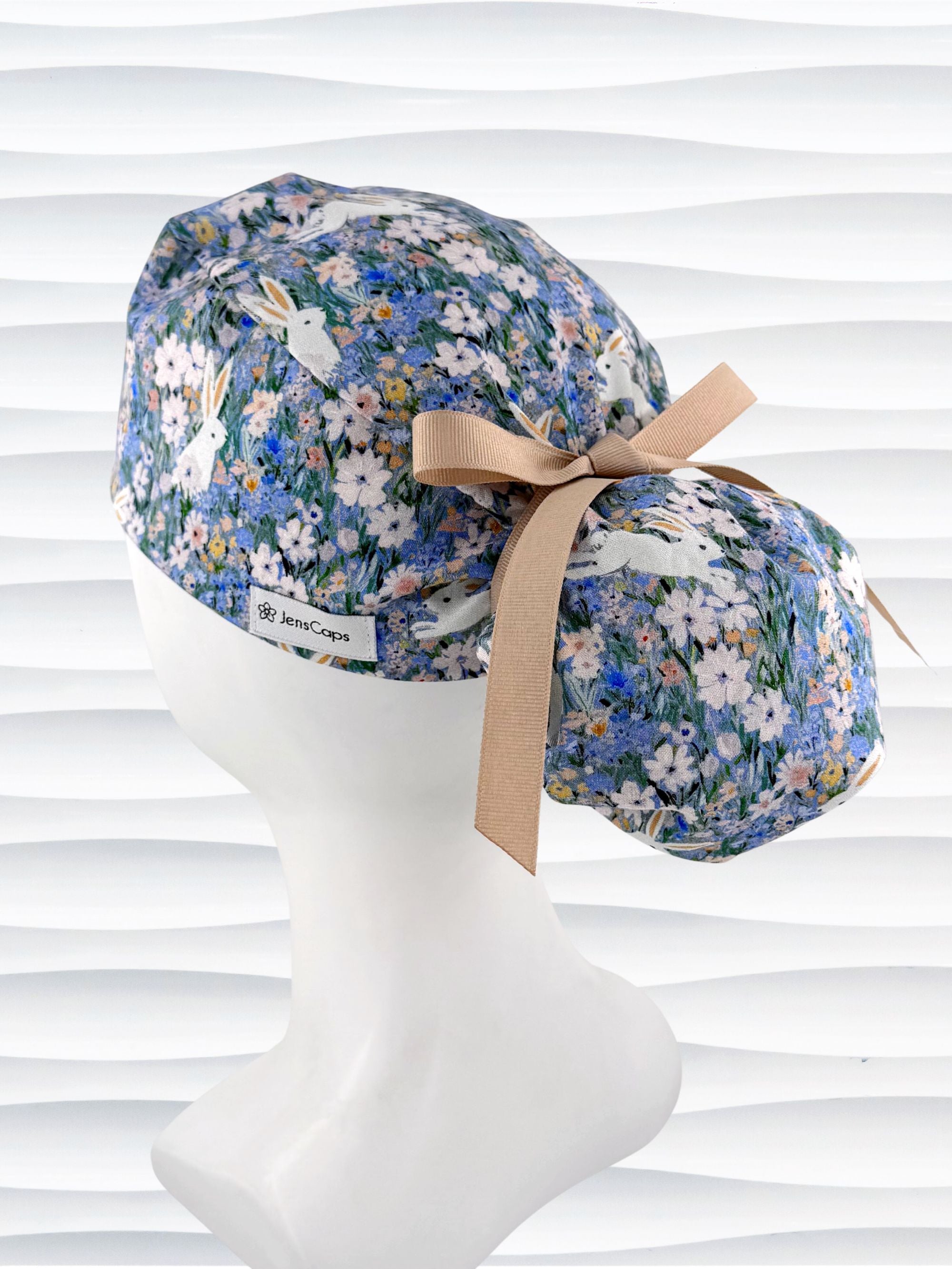 A ponytail style surgical scrub cap by JensCaps with white rabbits in various poses — sitting, leaping, and hopping — scattered across a dense meadow of wildflowers in periwinkle blue, soft pink, pale yellow, and green and finished with a ribbon tie and photographed on a mannequin head.