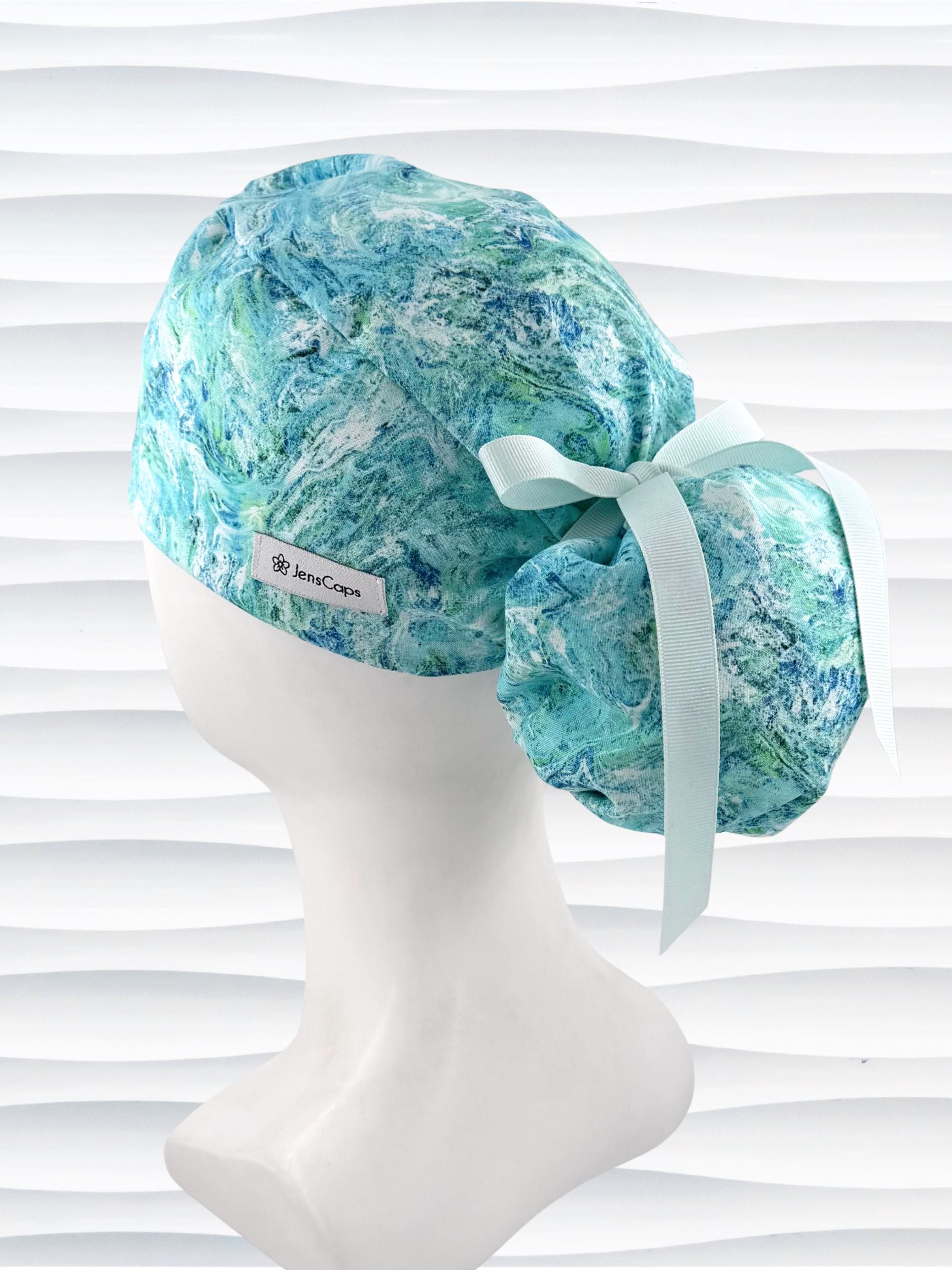 Ponytail style surgical scrub cap by JensCaps with abstract greens and blues on white cotton fabric and displayed on mannequin head.