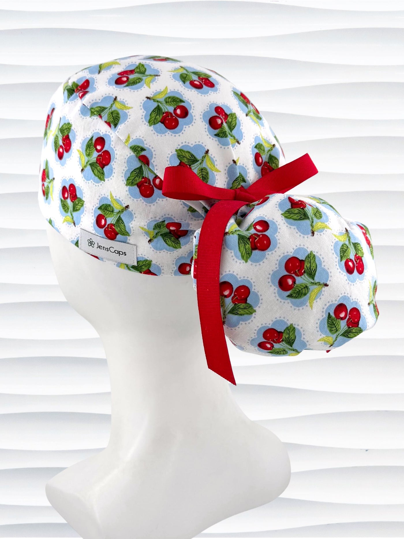 Ponytail surgical scrub cap by JensCaps with red cherries blue polka dots on white cotton fabric, finished with a ribbon tie, and diplayed on a mannequin head.