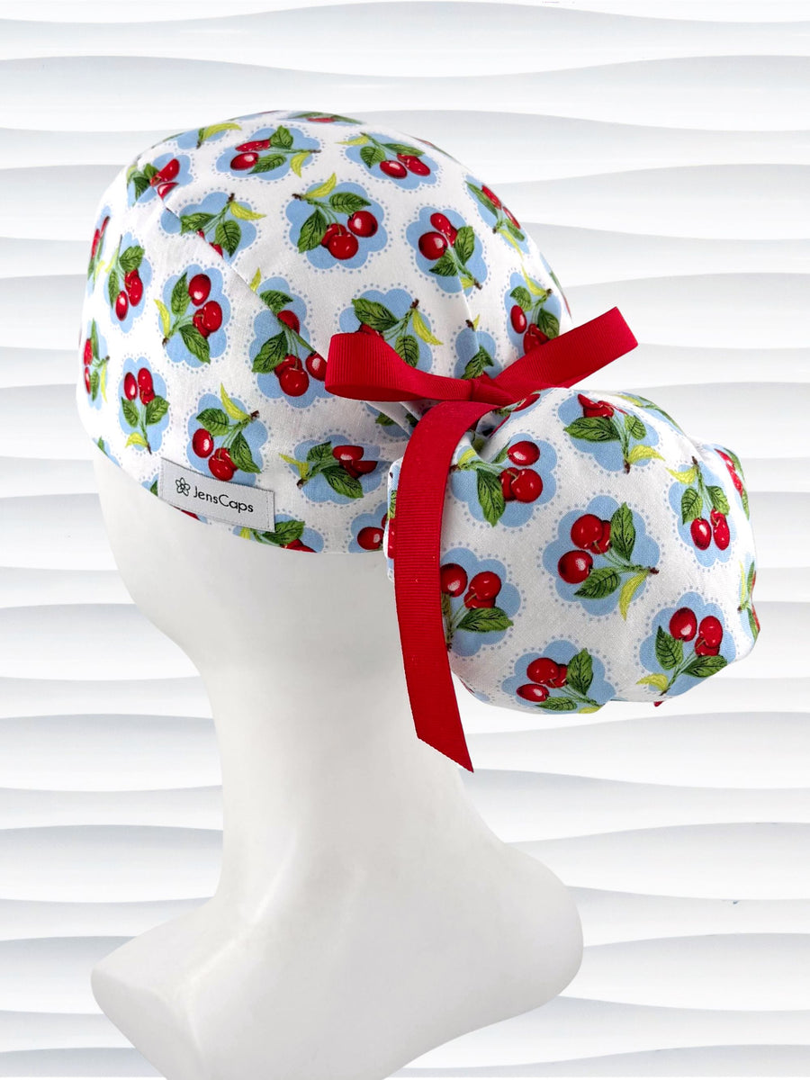 Ponytail surgical scrub cap by JensCaps with red cherries blue polka dots on white cotton fabric, finished with a ribbon tie, and diplayed on a mannequin head.