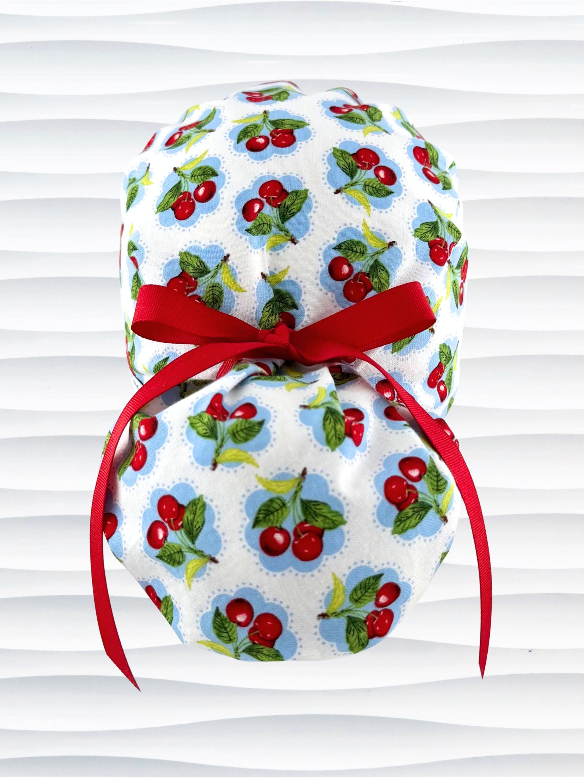 Ponytail surgical scrub cap by JensCaps with red cherries blue polka dots on white cotton fabric, finished with a ribbon tie, and diplayed on a mannequin head.