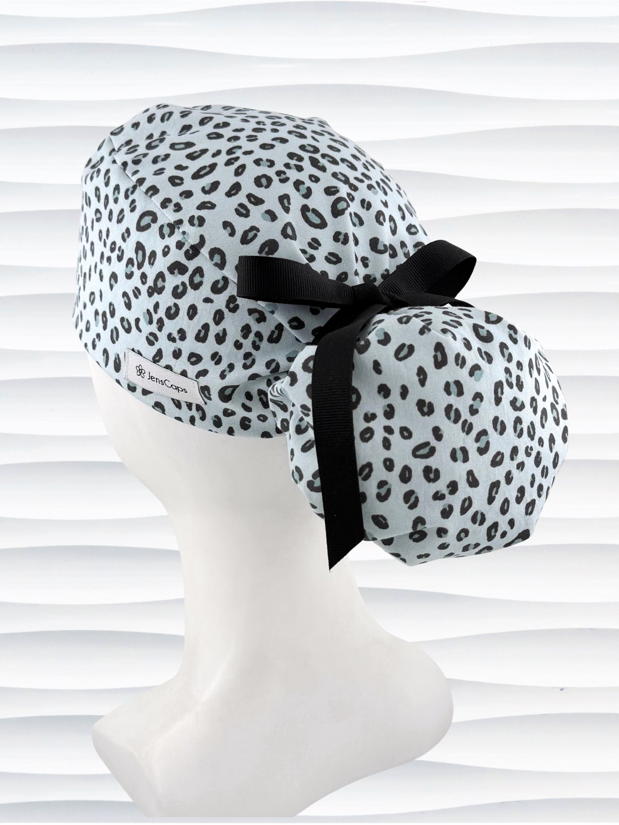 Ponytail style surgical scrub cap by JensCaps with a leopard print on light teal cotton fabric, tied with a ribbon, and displayed on a mannequin head.