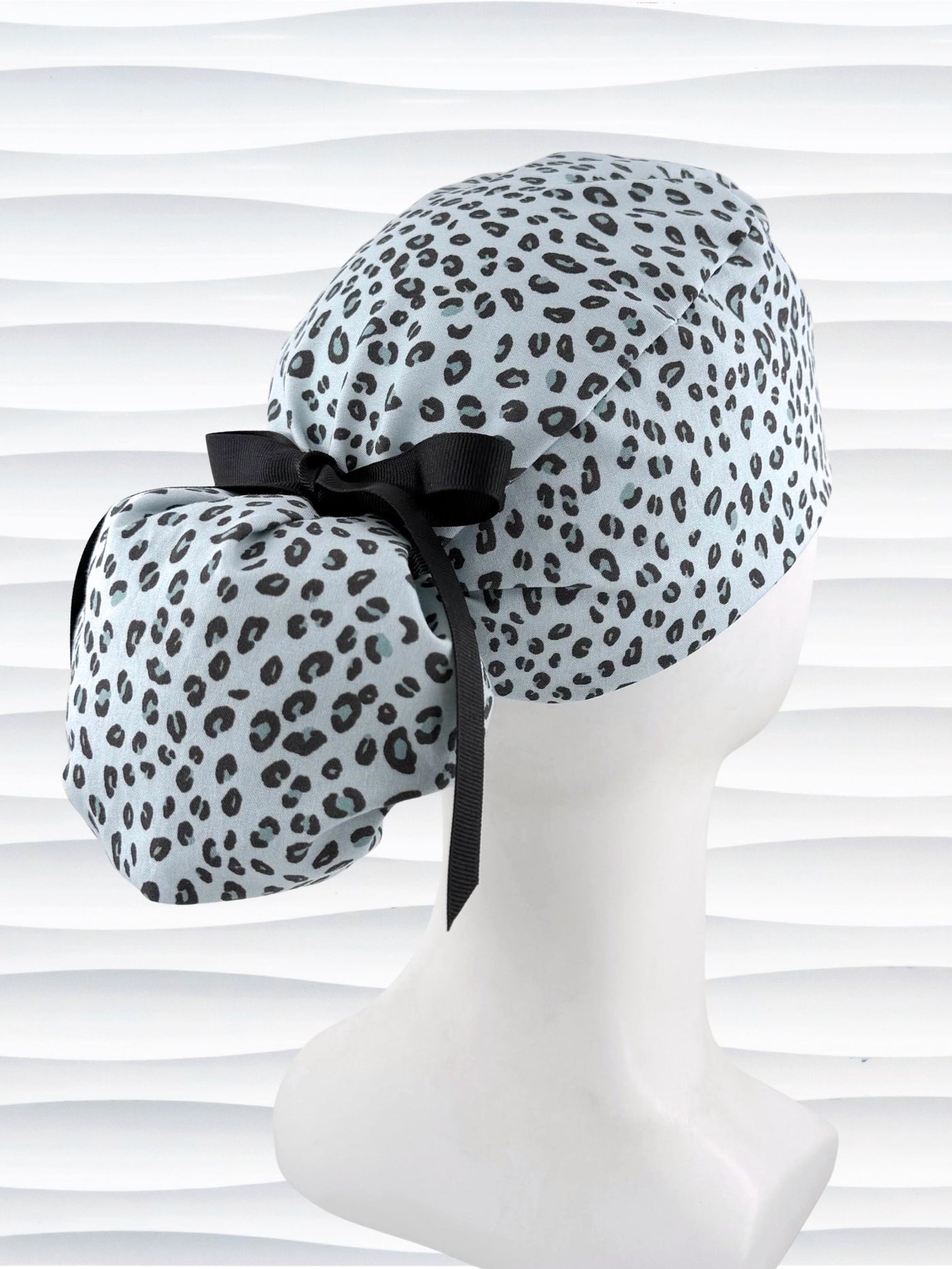Ponytail style surgical scrub cap by JensCaps with a leopard print on light teal cotton fabric, tied with a ribbon, and displayed on a mannequin head.