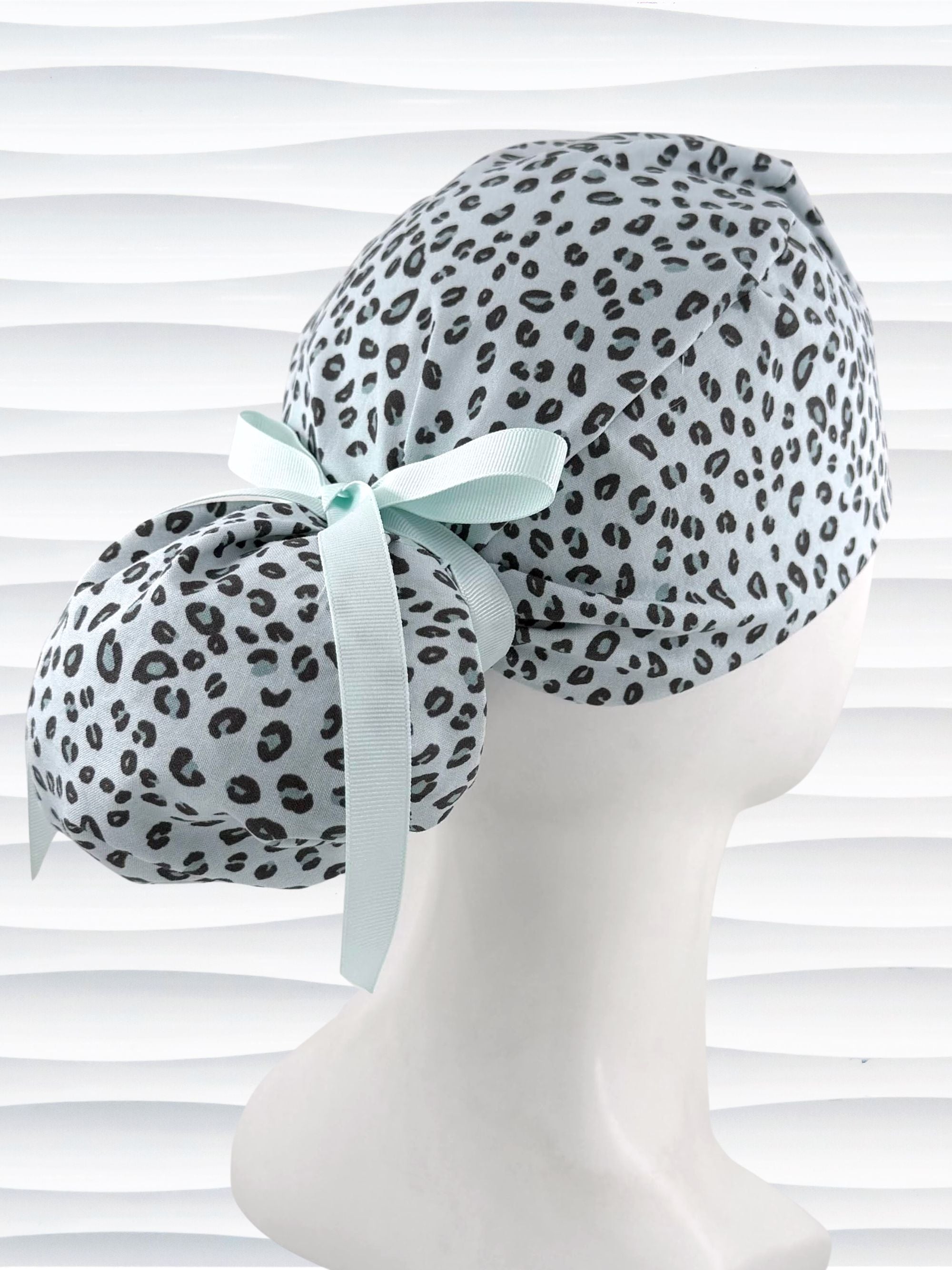 Ponytail style surgical scrub cap by JensCaps with a leopard print on light teal cotton fabric, tied with a ribbon, and displayed on a mannequin head.