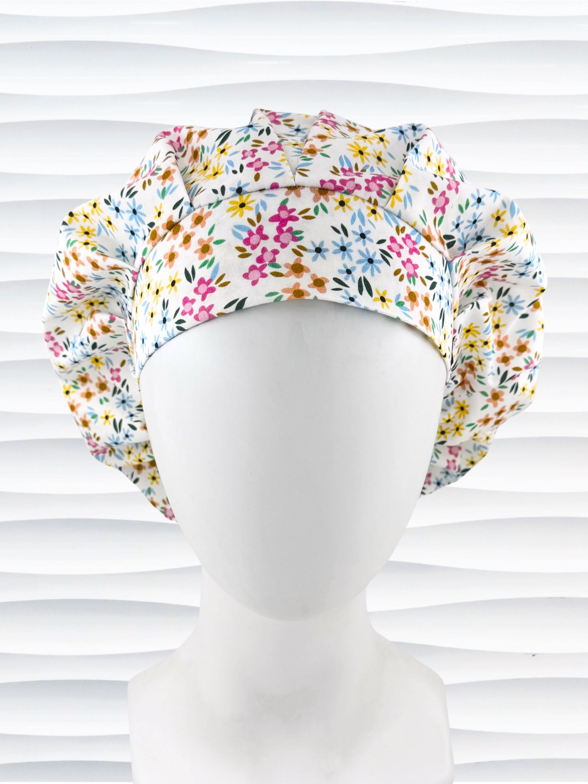 A white fabric bouffant style surgical cap by JensCaps with a colorful floral print, displayed on a white mannequin head against a white wavy background.