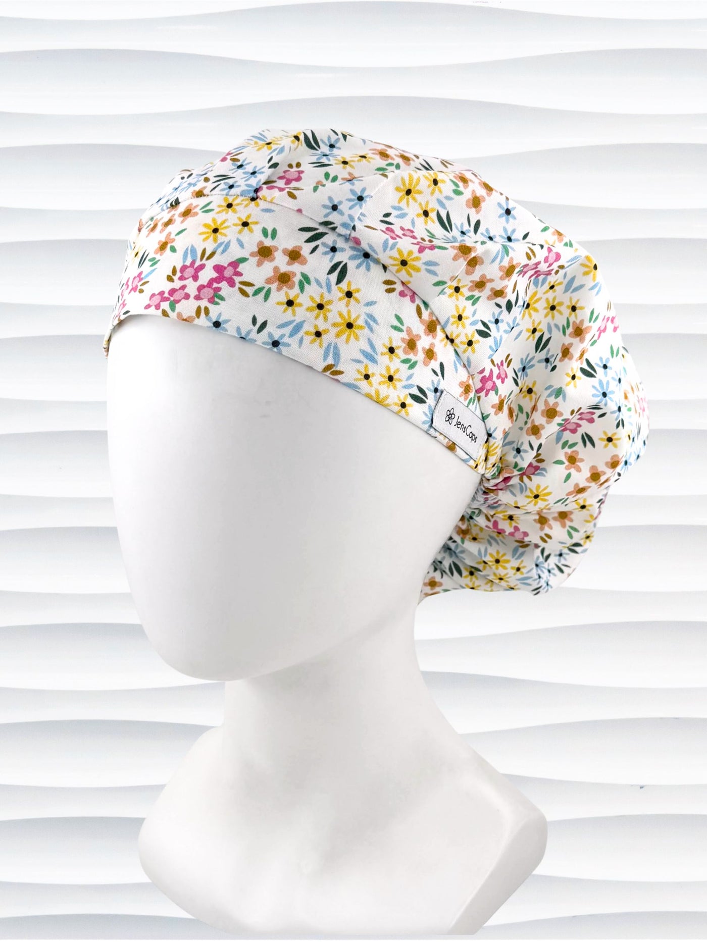 A white fabric bouffant style surgical cap by JensCaps with a colorful floral print, displayed on a white mannequin head against a white wavy background.