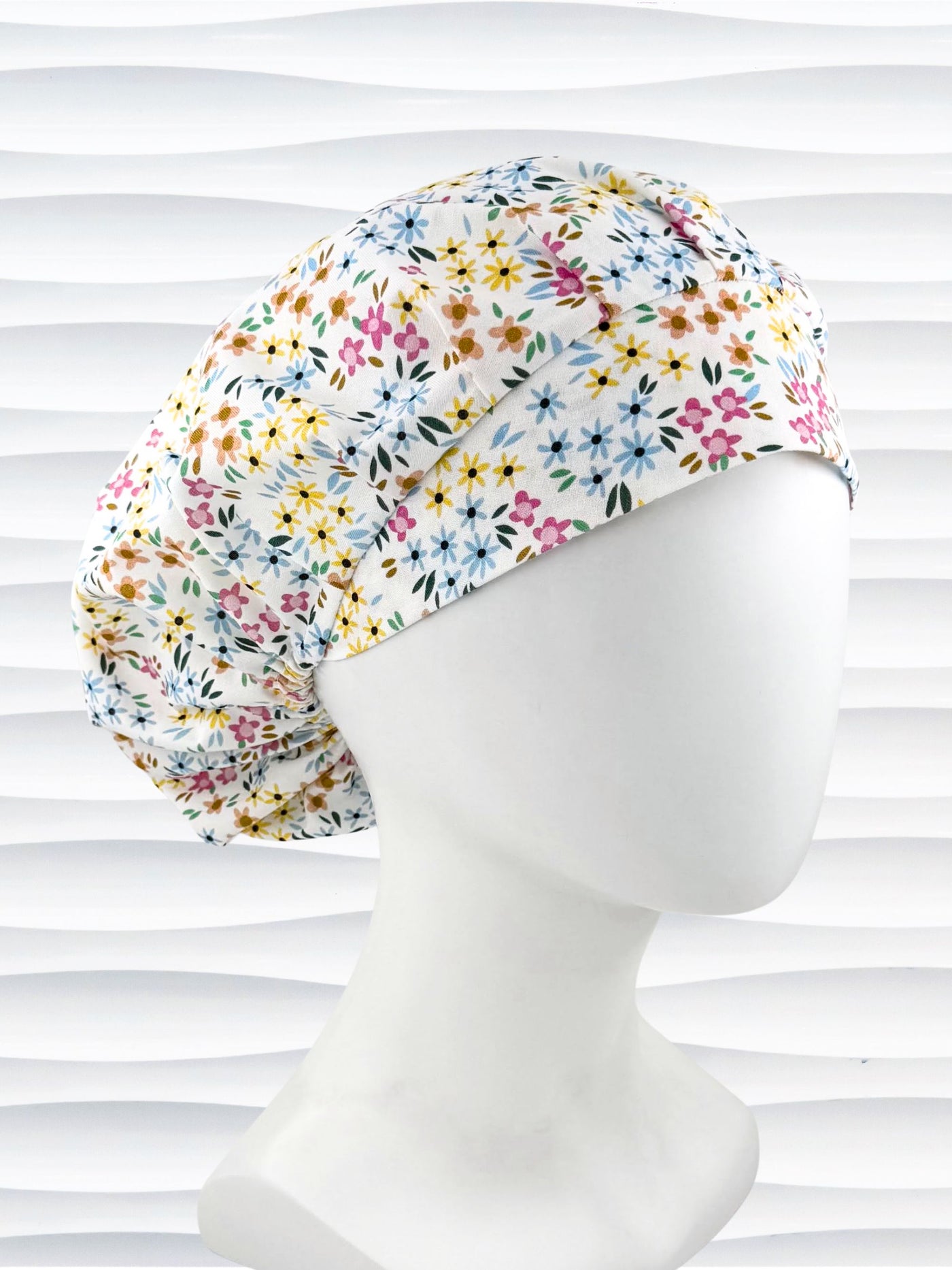 A white fabric bouffant style surgical cap by JensCaps with a colorful floral print, displayed on a white mannequin head against a white wavy background.
